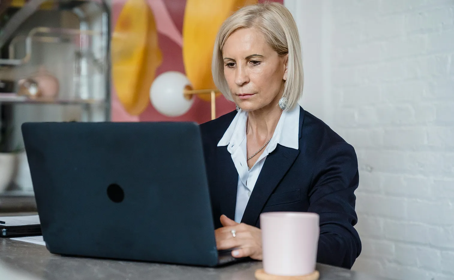 Focused middle-aged woman with short blonde hair working on a laptop at a desk with a mug in front of her.