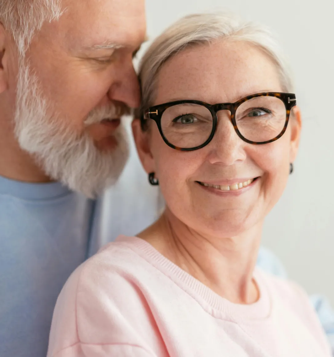 Smiling older woman with glasses in pink shirt with older man whispering to her.