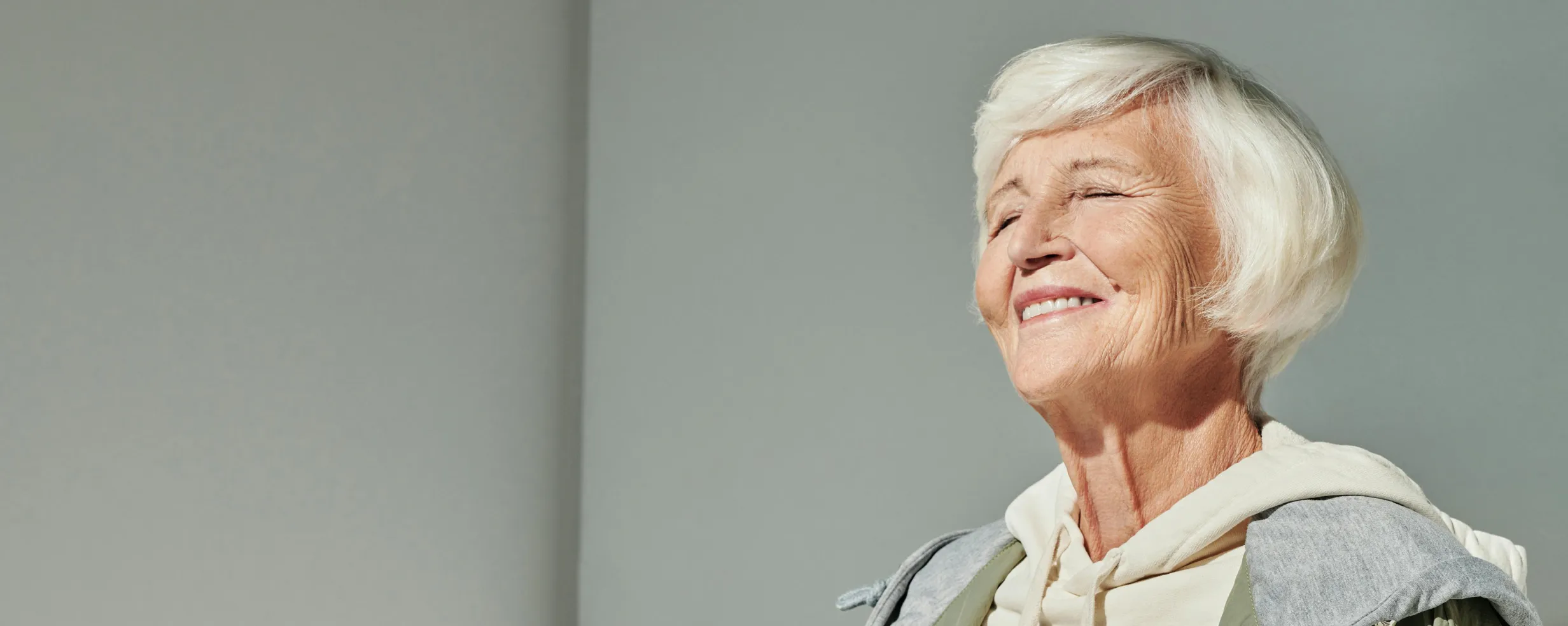 Smiling elderly woman with short white hair wearing a hoodie and jacket, eyes closed against a plain gray background.