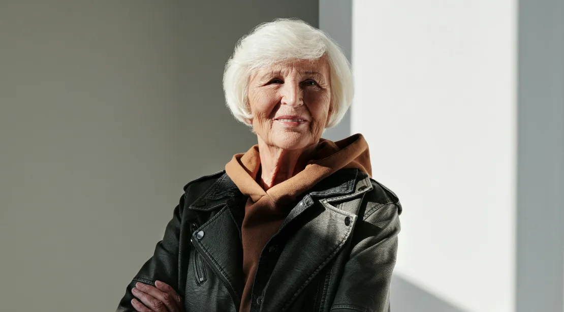 Smiling elderly woman with white hair wearing a brown hoodie and black leather jacket, standing with arms crossed.