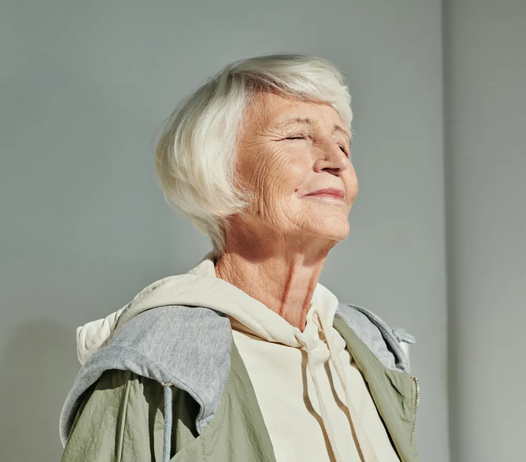 Smiling elderly woman with short white hair, wearing a cream hoodie and green jacket, eyes closed appreciating sunlight.