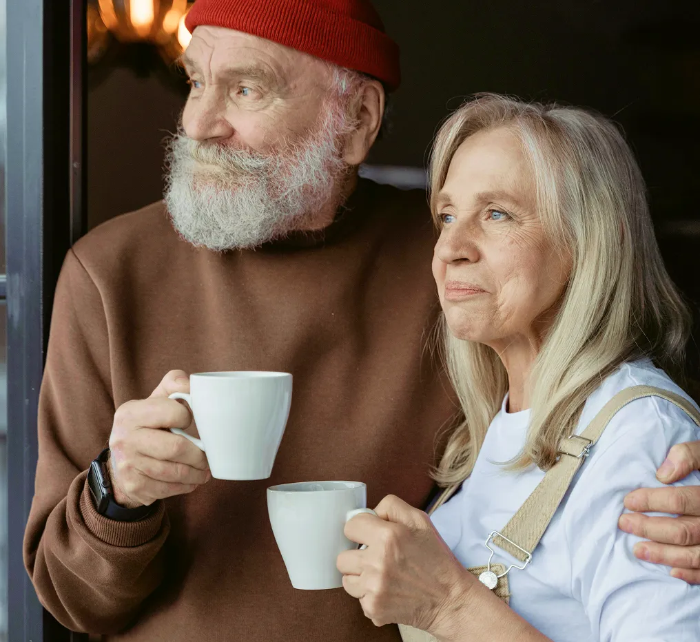 Older couple standing by a window, holding white mugs and looking outside thoughtfully.