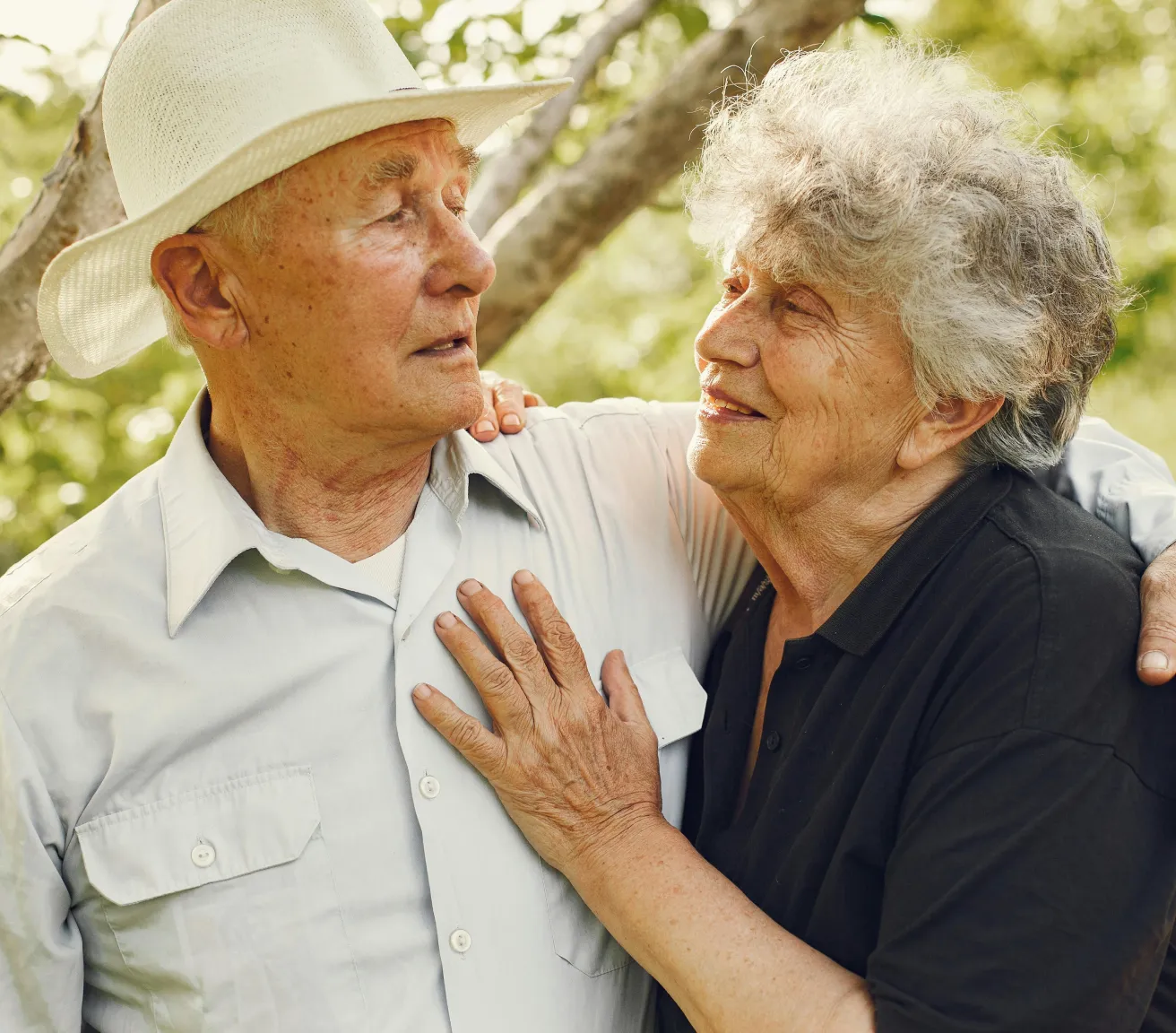 Elderly couple outdoors gazing at each other warmly with the woman’s hand on the man’s chest and his arm around her.