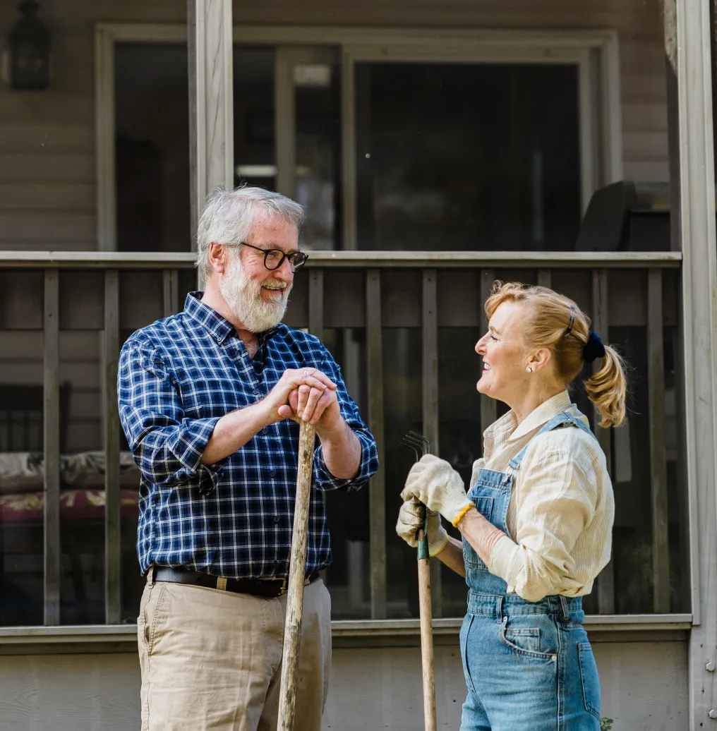 Smiling elderly man and woman standing outdoors with gardening tools, engaged in conversation.