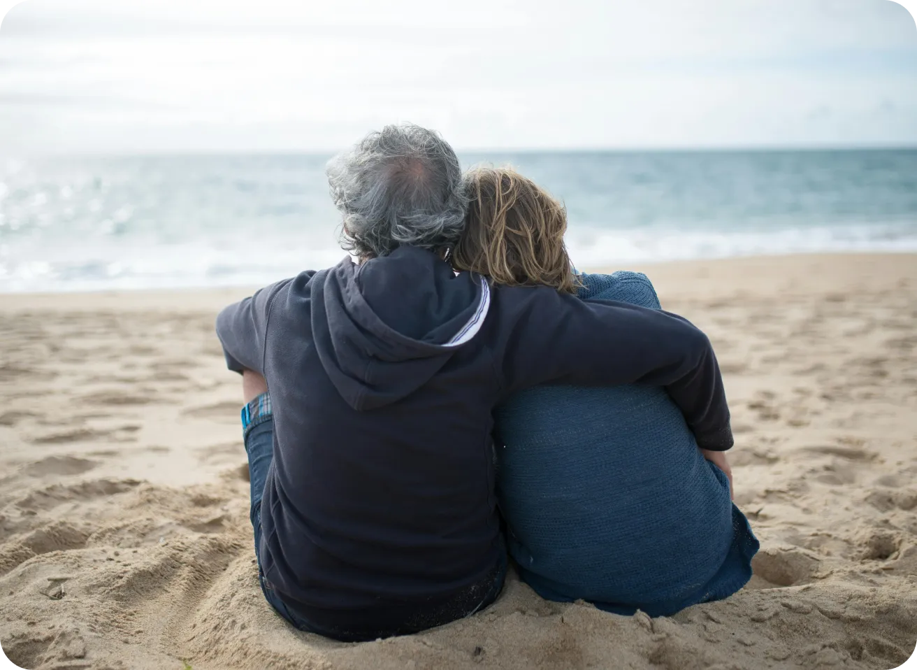 Couple sitting closely together on sandy beach facing the ocean, with one person's arm around the other.