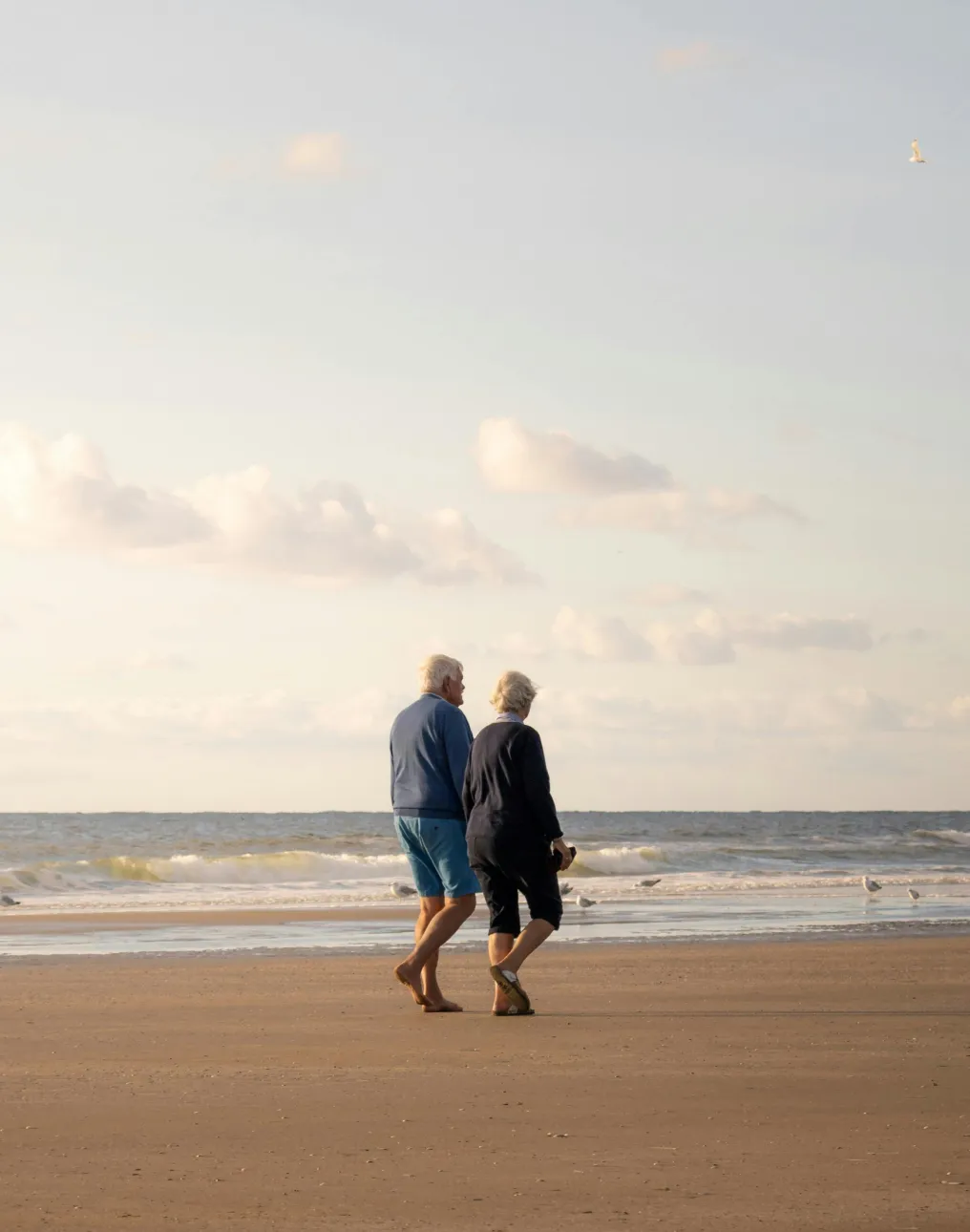 Elderly couple walking barefoot on a sandy beach near the ocean under a cloudy sky.