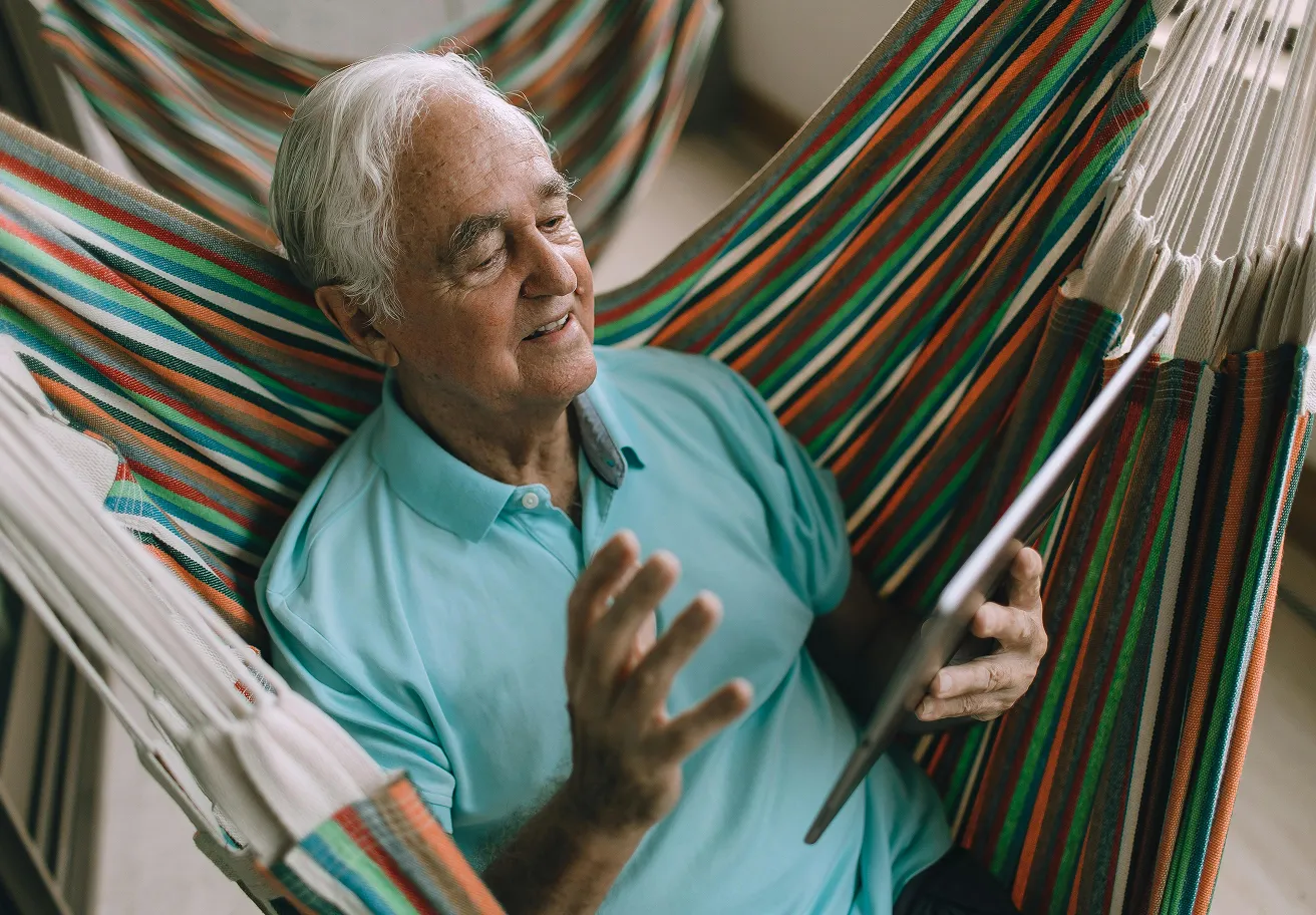 Elderly man in a blue shirt sitting in a colorful striped hammock using a tablet.