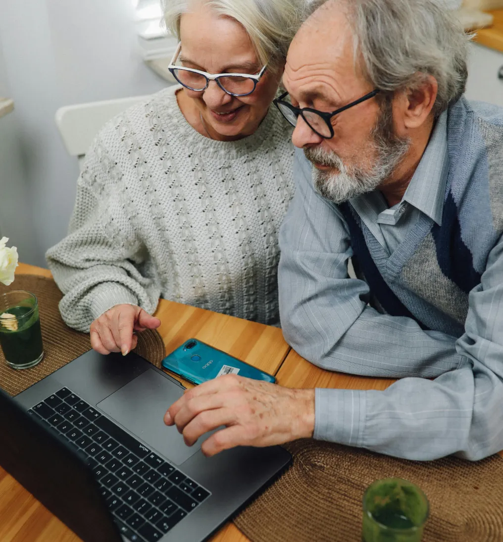 Older couple sitting at a table using a laptop together, with a smartphone and drinks nearby.