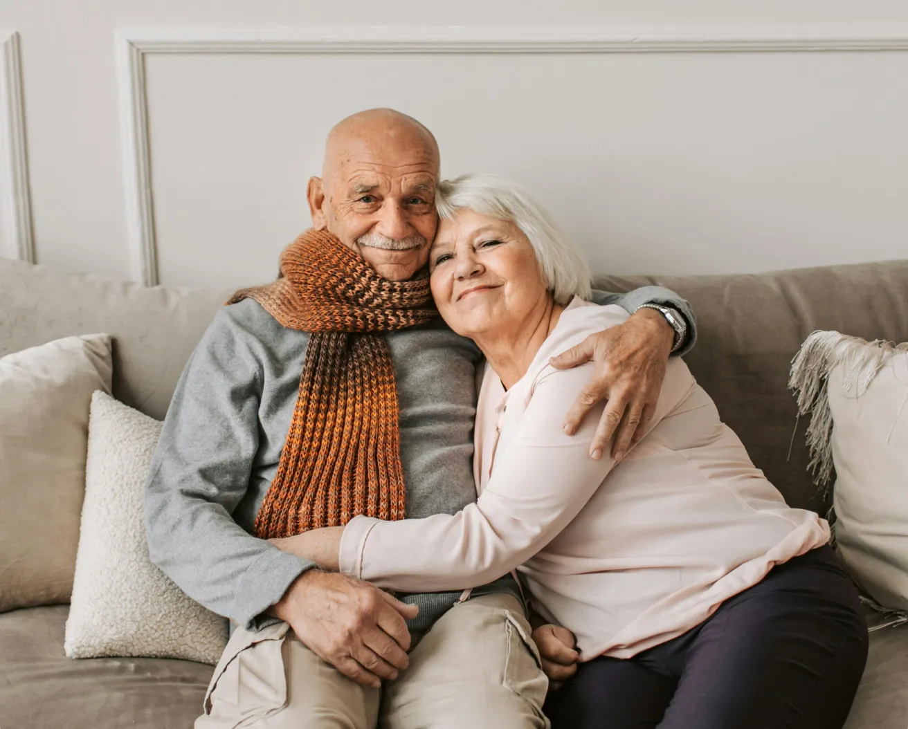 Elderly couple sitting on a couch, smiling and hugging each other warmly.