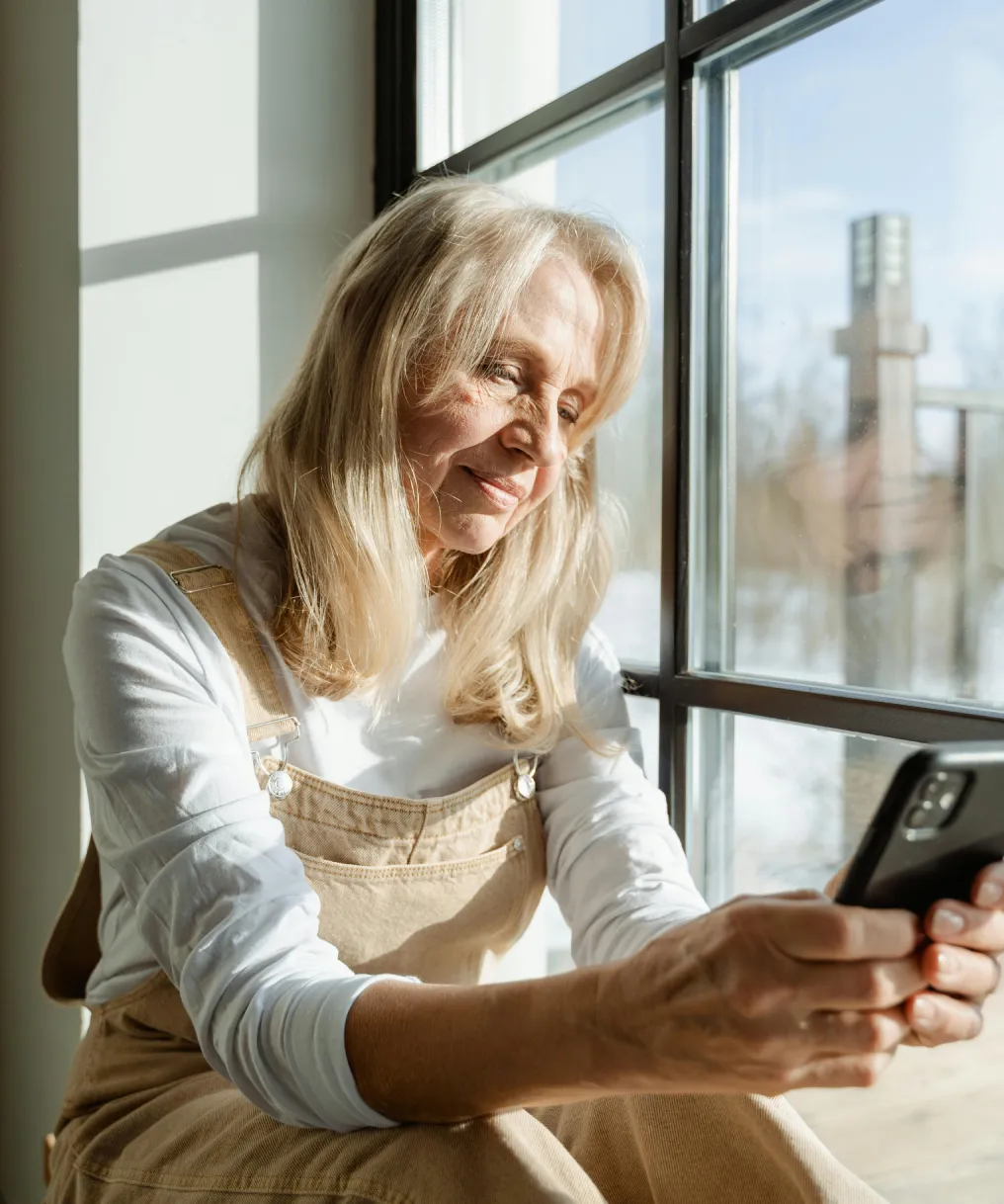 Smiling mature woman sitting by a window using a smartphone, with sunlight on her face.
