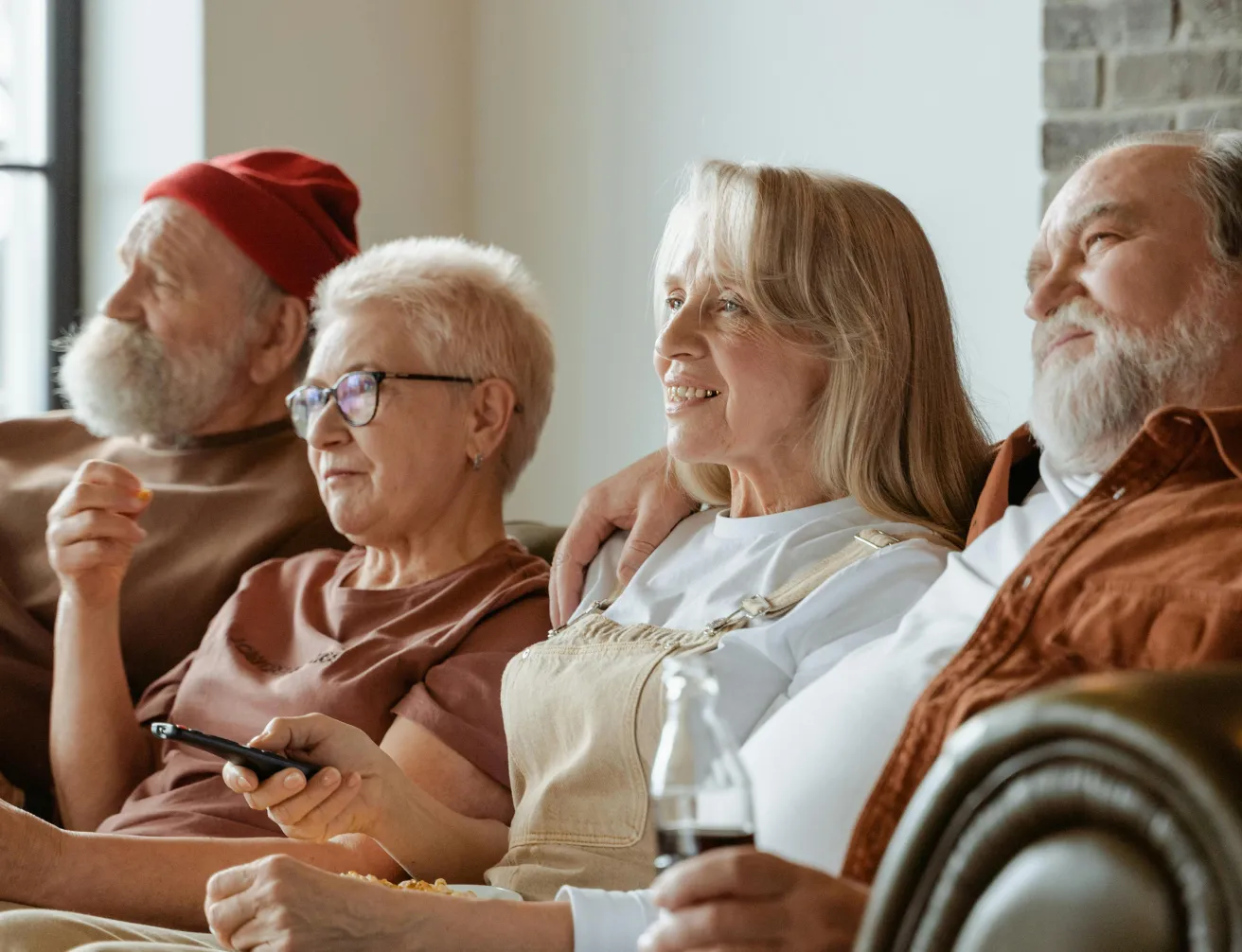 Four elderly people sitting closely on a couch, watching TV together, with one holding a remote and another holding a beverage bottle.
