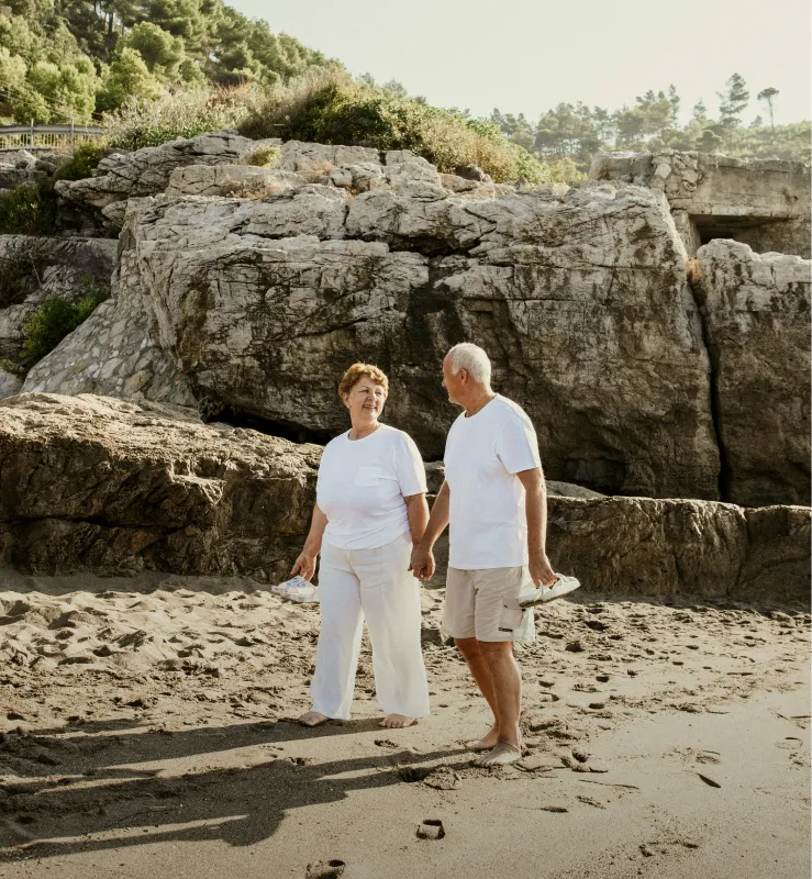 Elderly couple holding hands and walking barefoot on a sandy beach near rocky cliffs, both wearing white shirts.