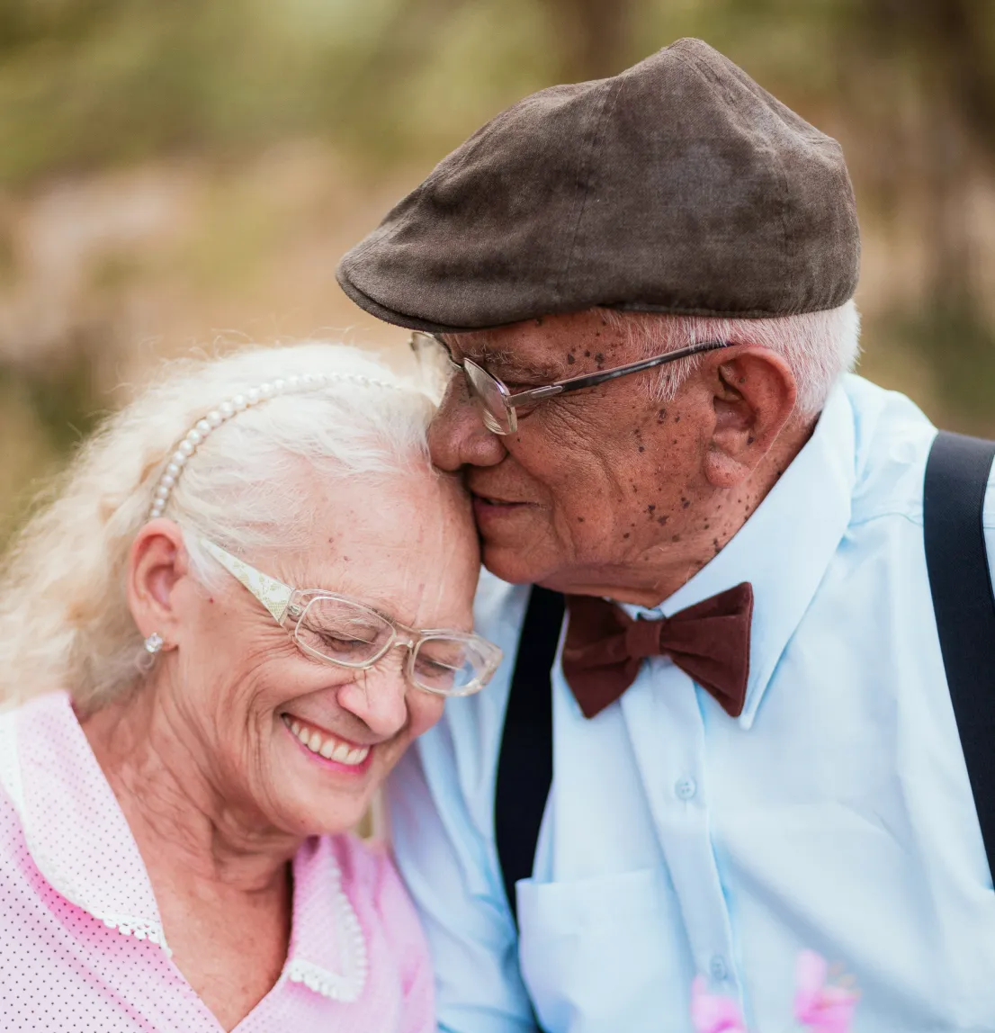 Elderly man in a brown flat cap and bow tie gently resting his forehead against a smiling elderly woman with glasses and a pearl headband.