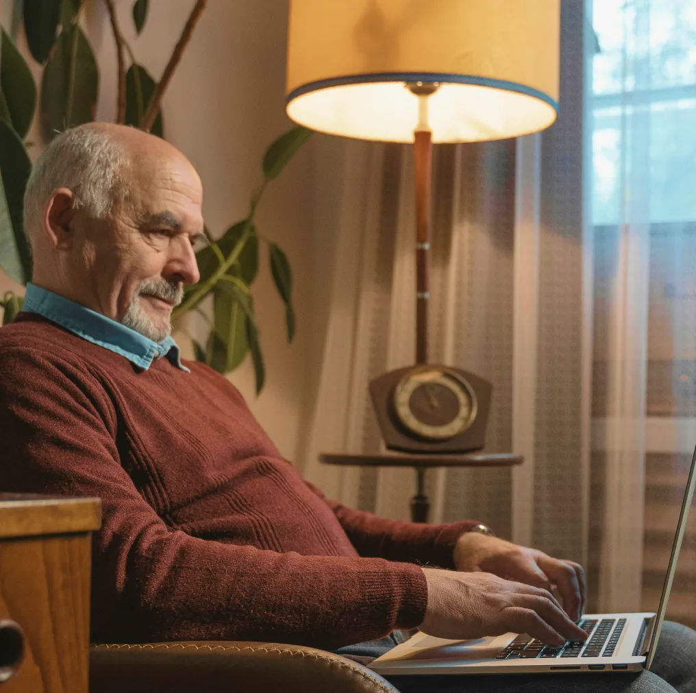 Elderly man in a red sweater sitting in a chair using a laptop with a lit lamp and plant in the background.