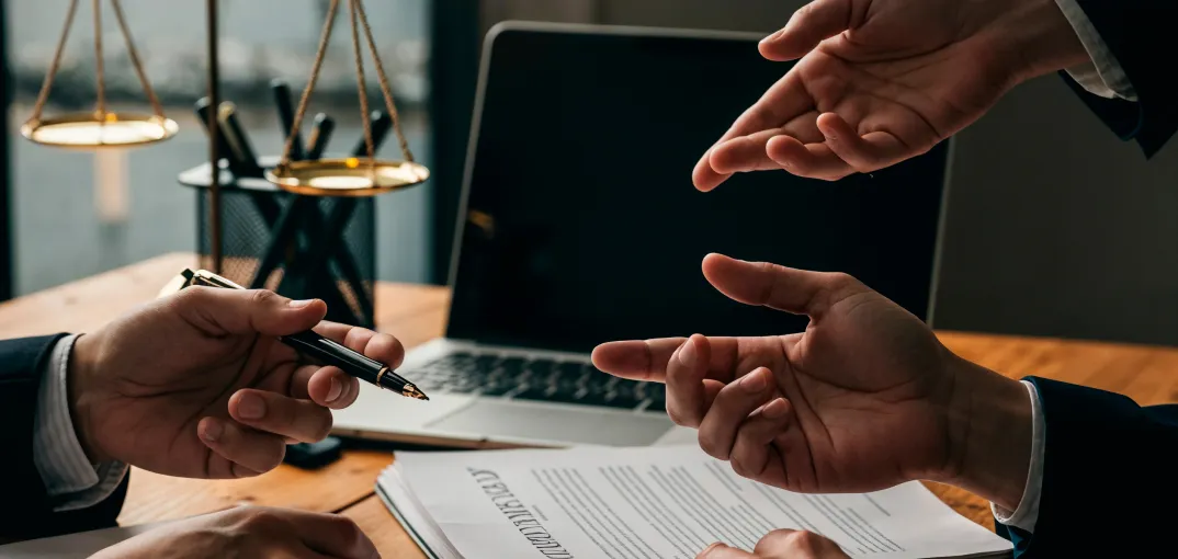 Two people in a meeting discussing a legal document with a laptop and balance scale on a wooden desk.