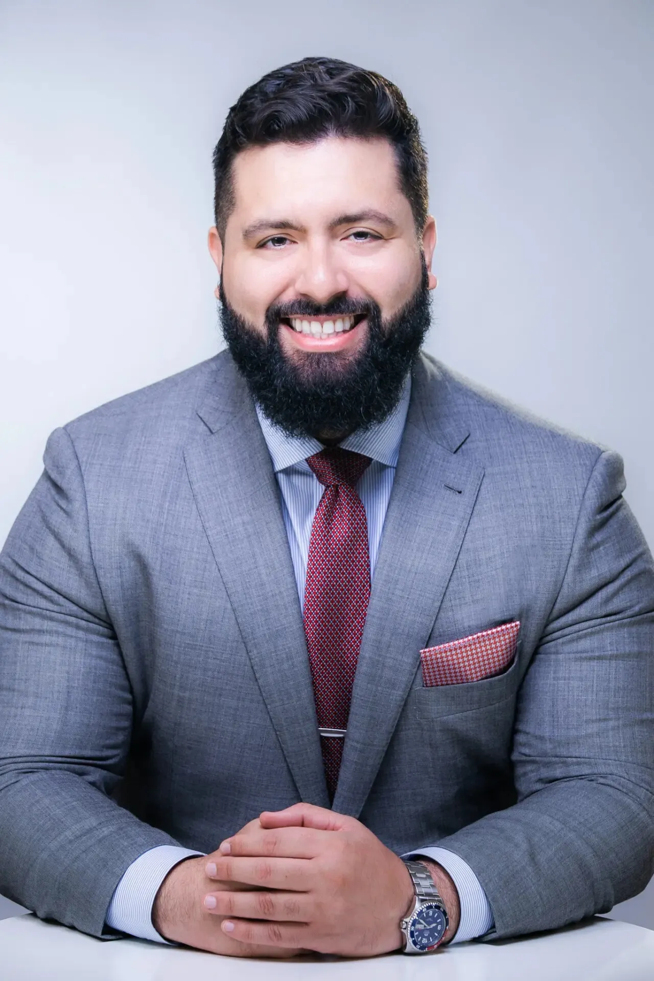 Smiling man with dark hair and beard wearing a gray suit, red patterned tie, and watch, seated with hands clasped.