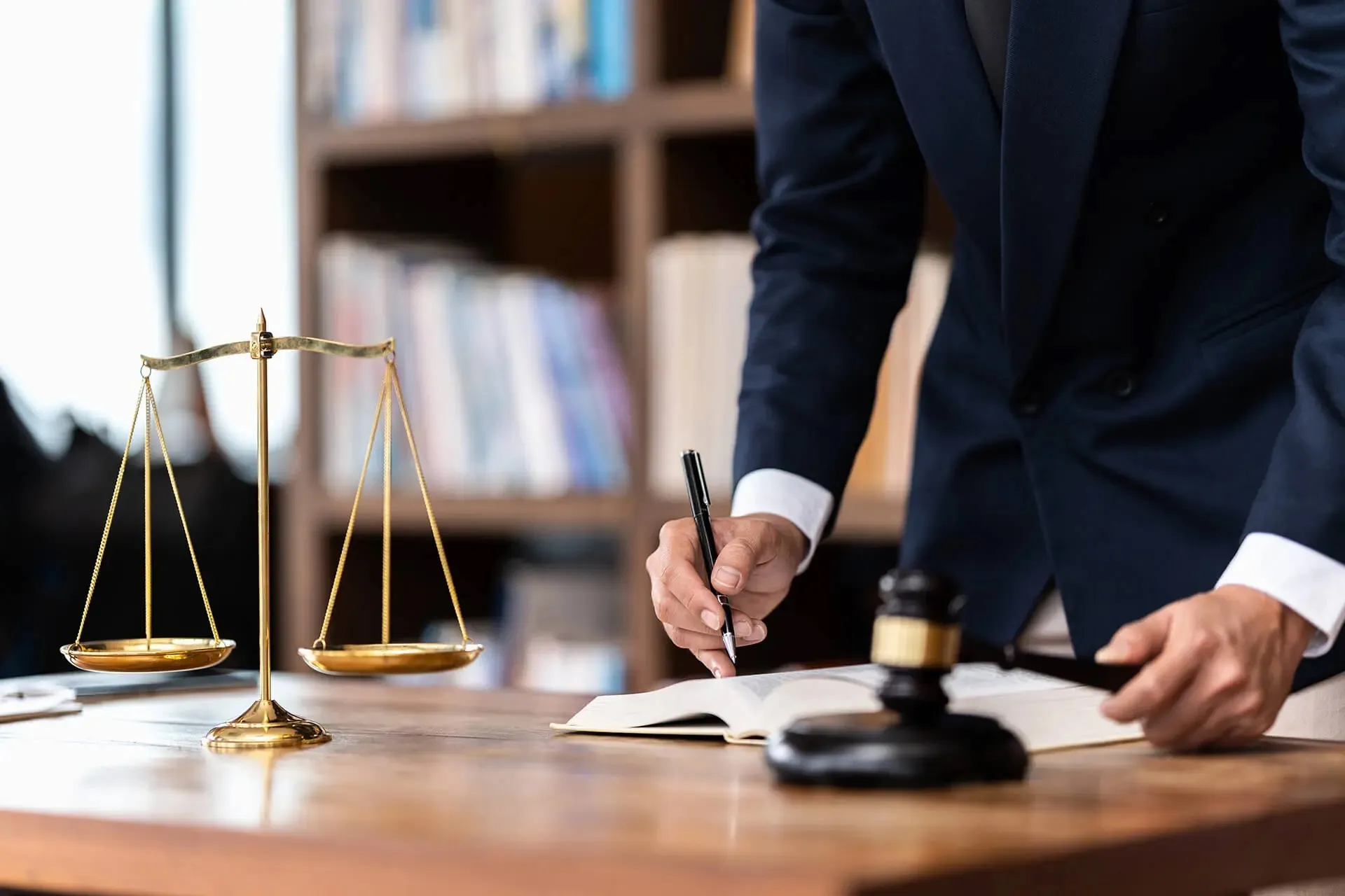 Person in navy suit writing in an open book with a gavel and gold balance scale on a wooden table.