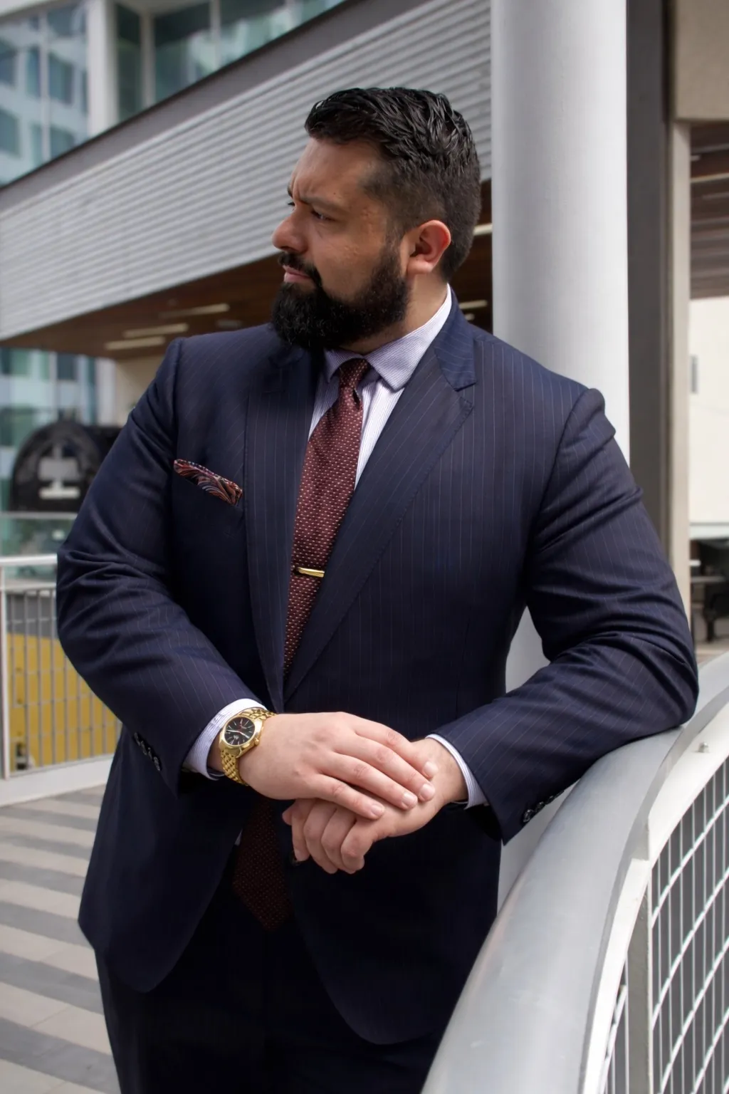 Man with beard and dark hair in a navy pinstripe suit, red tie, and gold watch, leaning on a railing outdoors.