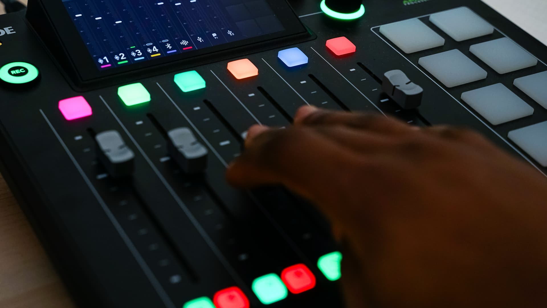 Hand adjusting sliders on a sound mixer with colorful illuminated buttons and a screen above.