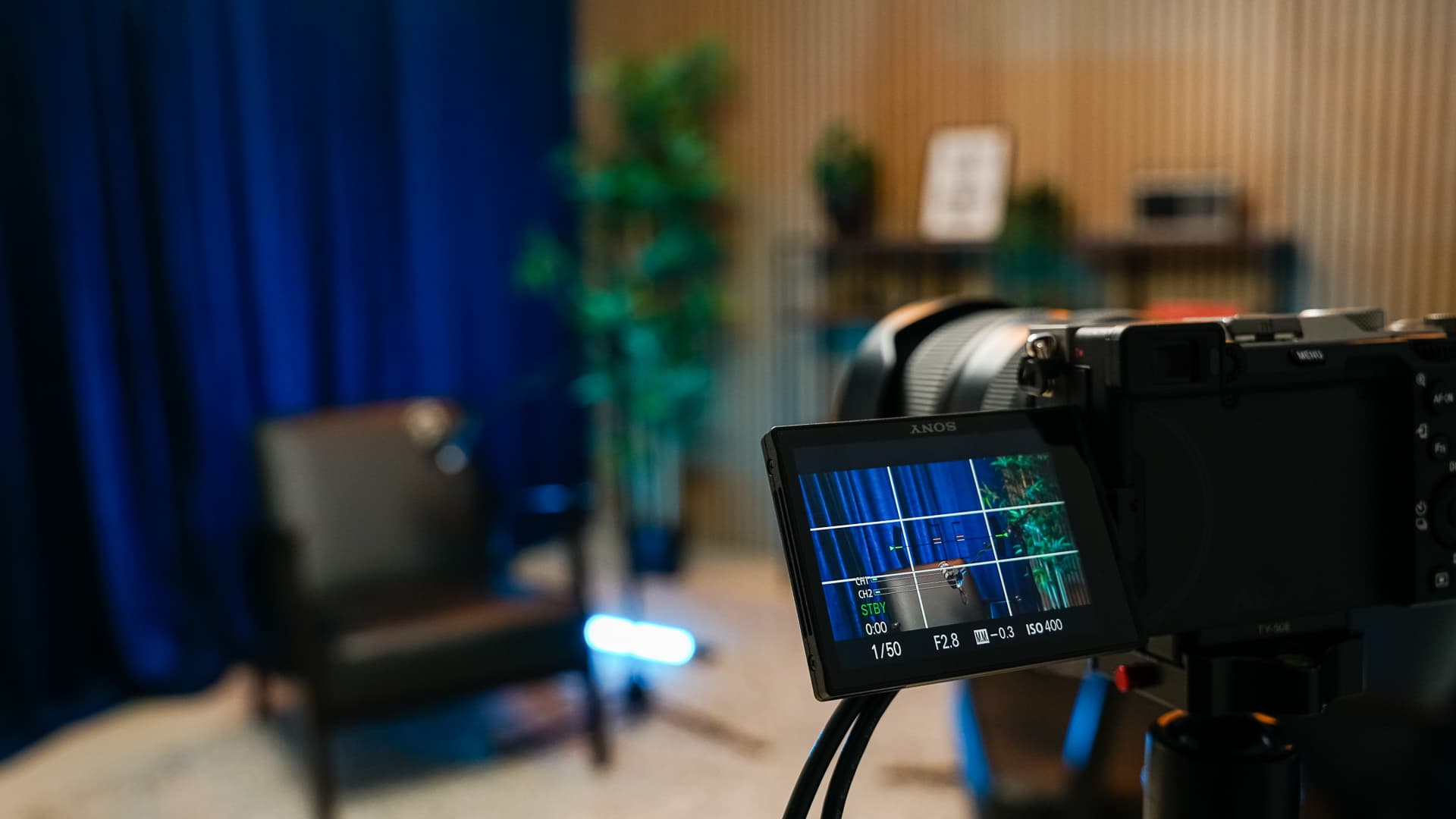 Camera monitor displays a chair and plants in a recording studio with blue curtains and wooden walls.