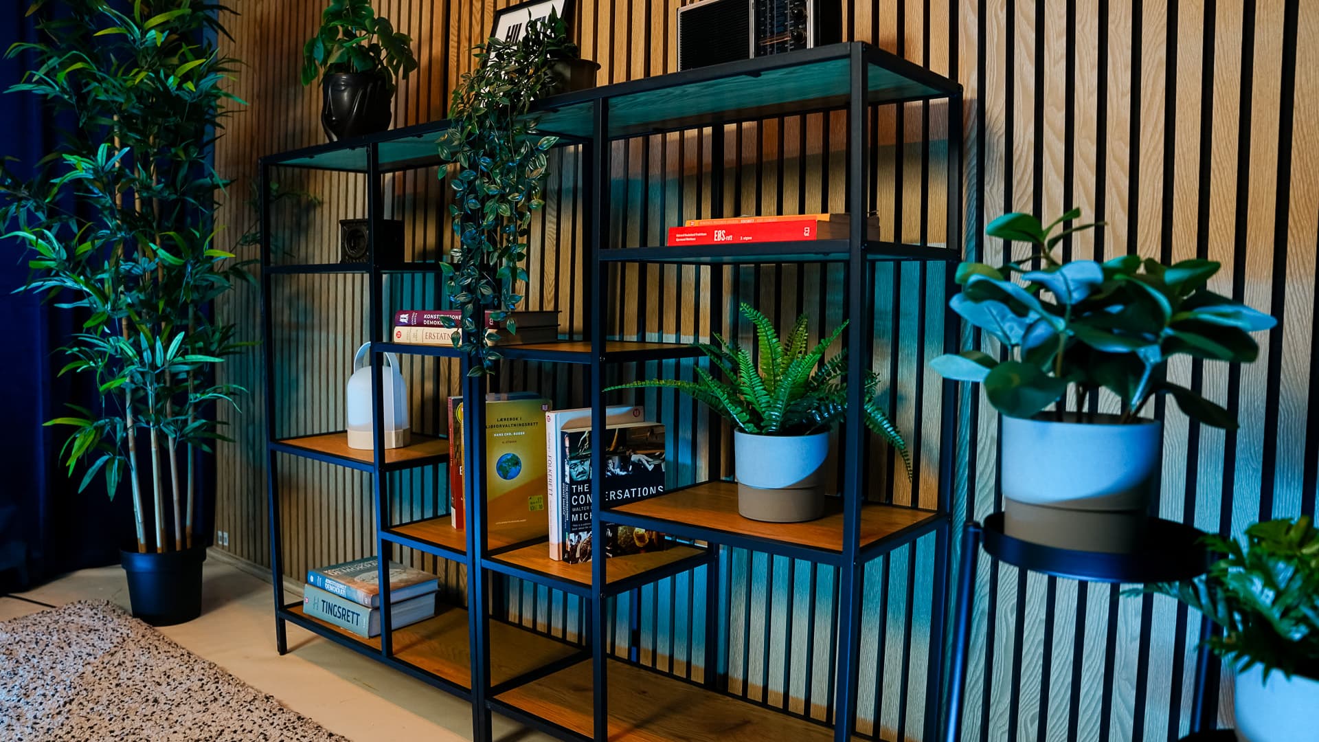 Black metal and wood shelf with books and plants against a wooden slat wall next to a tall potted plant.