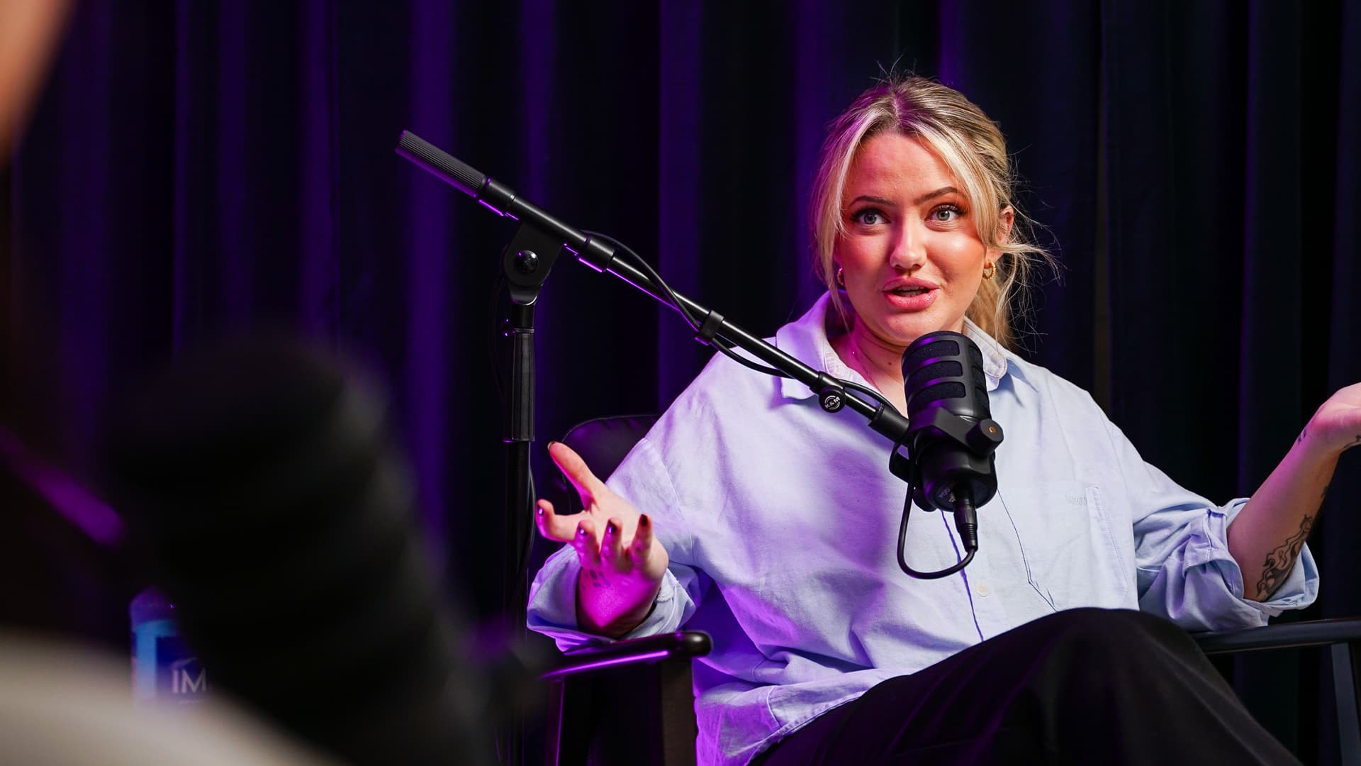 Woman with blonde hair speaking into a microphone during a podcast recording in front of black curtains.