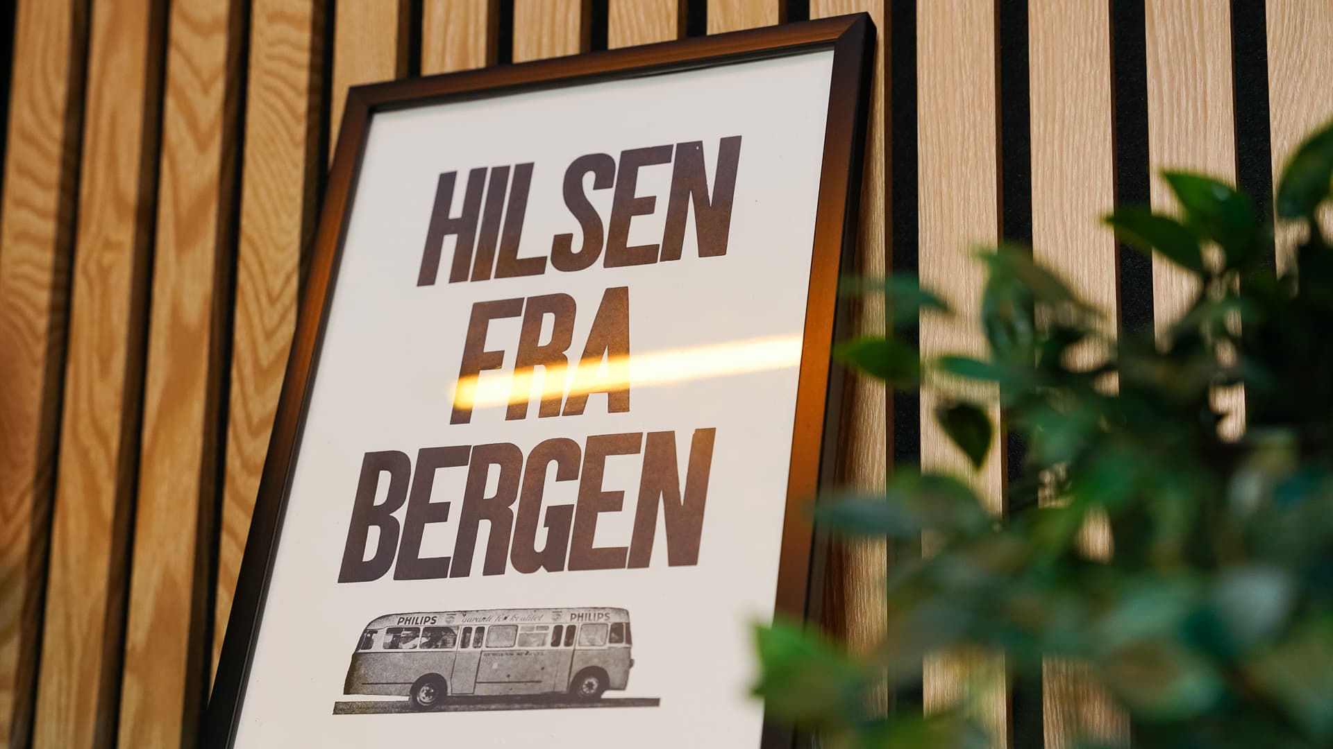 Framed sign with 'Hilsen fra Bergen' text and a vintage bus illustration on wood panel wall.