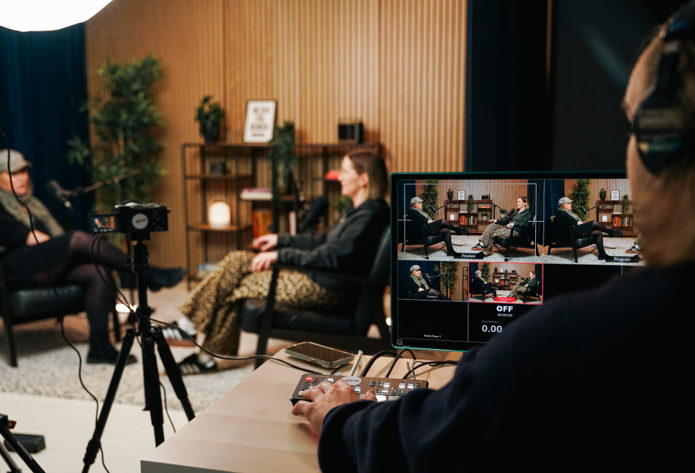 Person controlling livestream video showing two women seated and talking in a cozy studio setup.