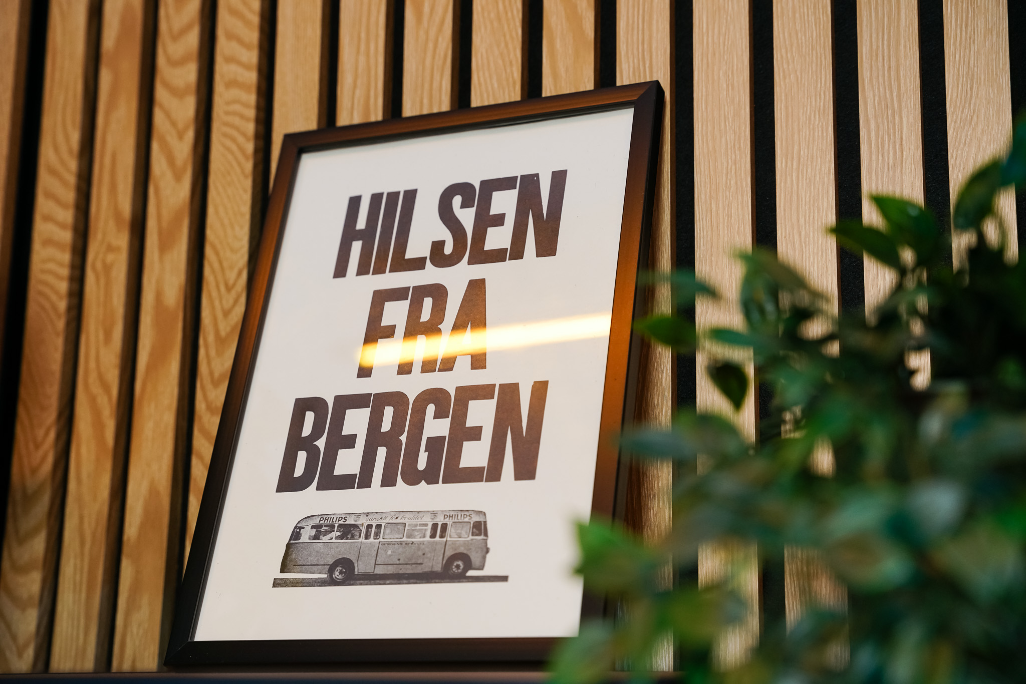 Framed sign with bold text 'Hilsen fra Bergen' and a vintage bus illustration leaning against a wooden wall.