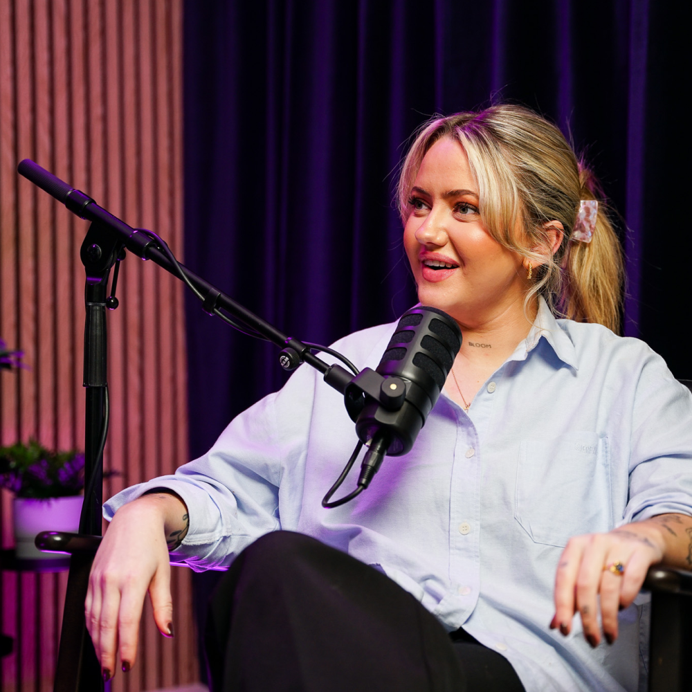 Woman with blonde hair in a ponytail speaking into a microphone during a podcast recording.