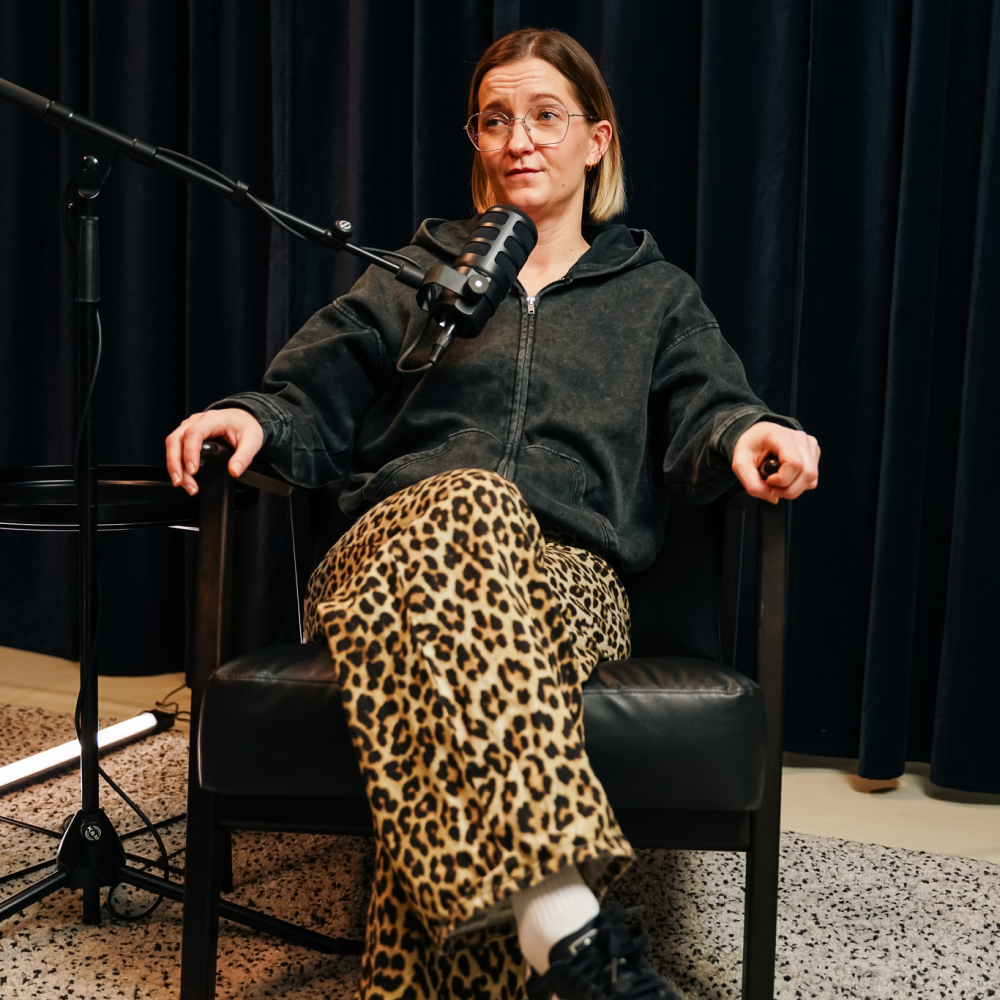 Person with glasses in a black hoodie and leopard pants sitting with crossed legs near a microphone.