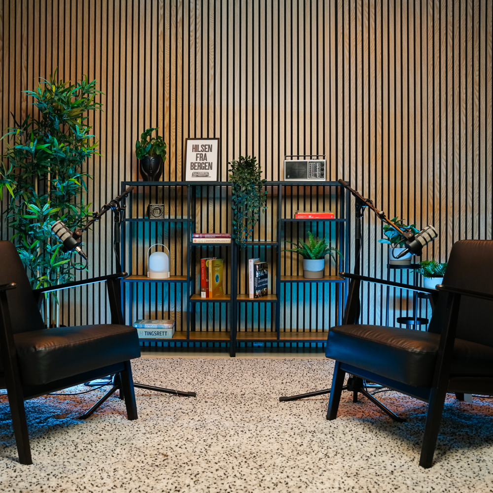 Two black chairs with microphones face each other in a podcast studio with plants and shelves.