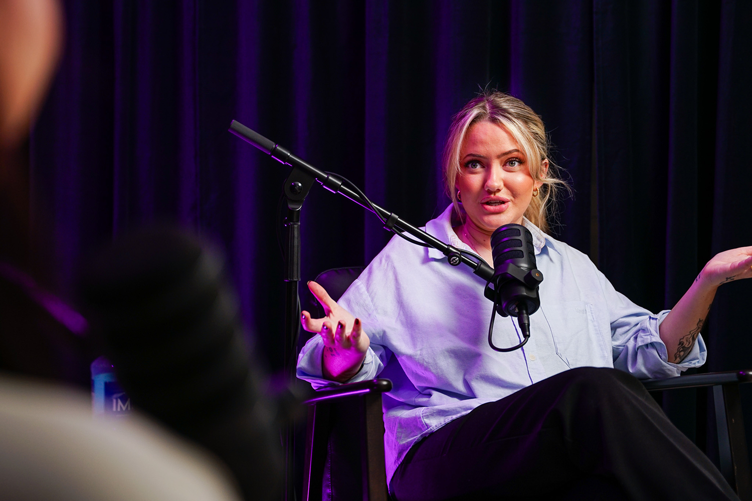 Blonde woman with light blue shirt speaks into microphone during a recording with dark purple curtain background.