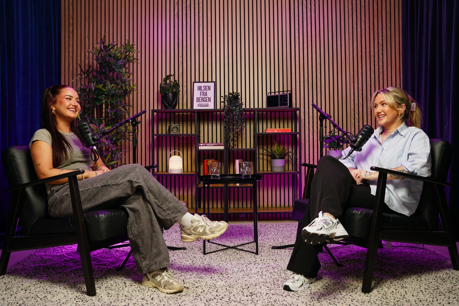 Two women smiling and talking in a podcast studio with microphones and colorful lighting.
