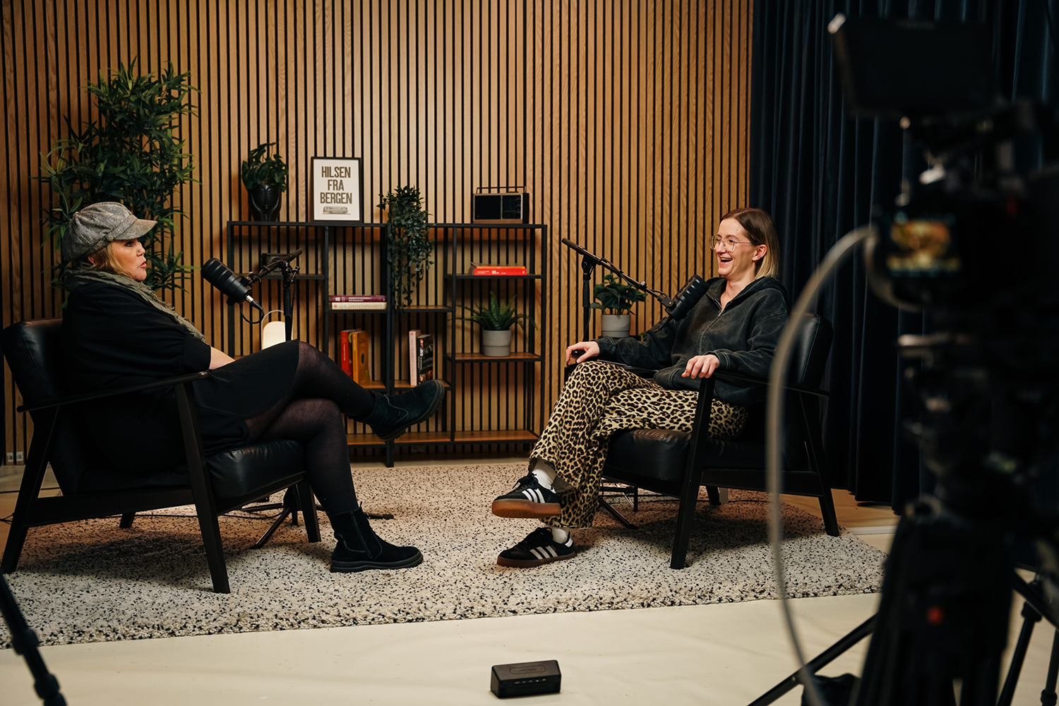 Two women seated with microphones having a podcast conversation in a cozy studio with wooden wall.