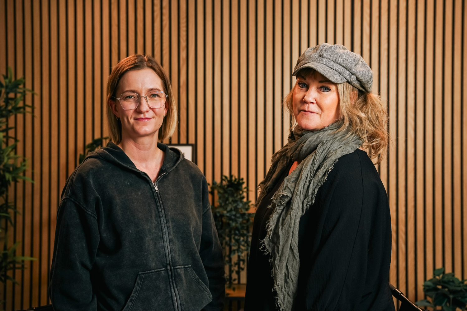 Two women standing in front of wooden slat wall, one wearing glasses and hoodie, the other a hat and scarf.