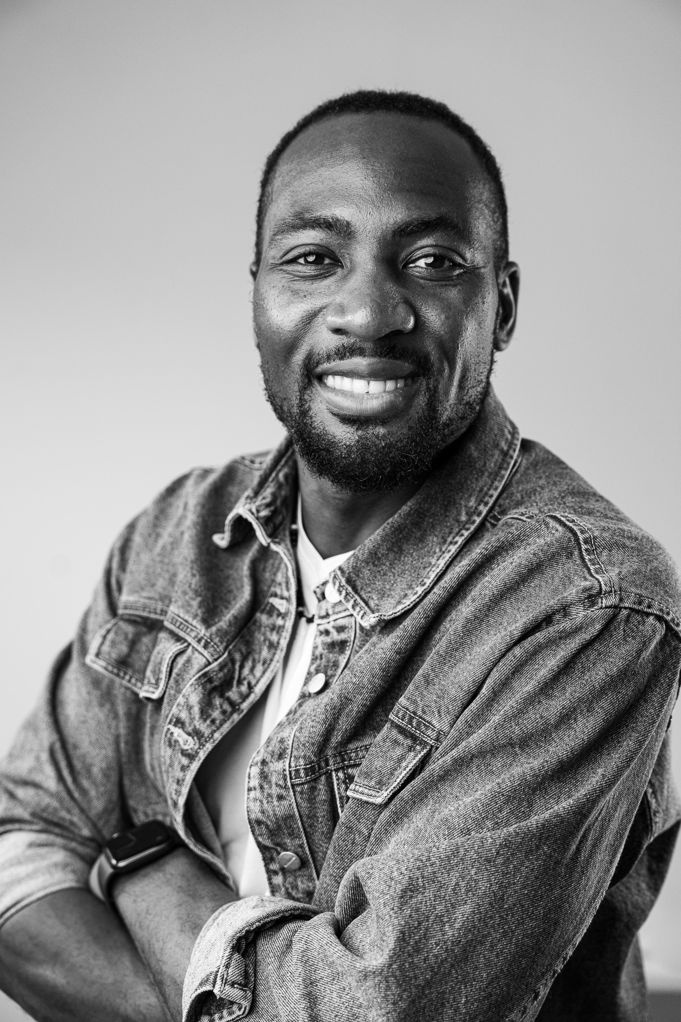 Smiling man with beard wearing a denim jacket and crossed arms in a black and white photo.