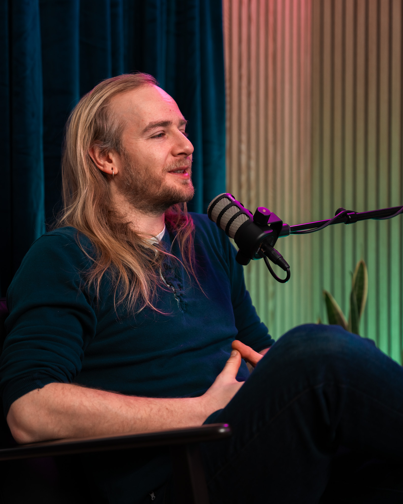 Man with long hair in a dark shirt speaking into a microphone while seated in a studio.