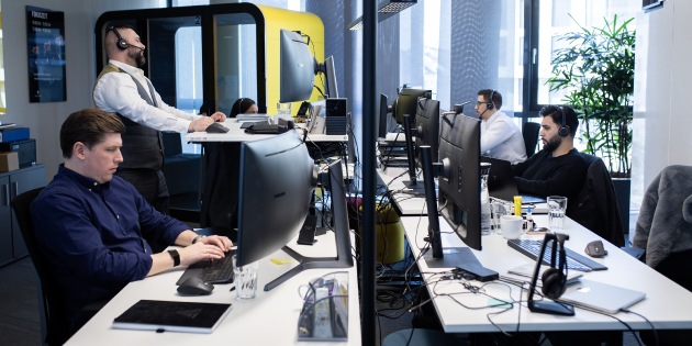 Vier Personen sitzen oder stehen an Schreibtischen und arbeiten konzentriert