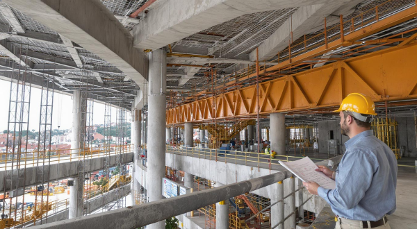 Ein Ingenieur steht auf einer Baustelle mit sichtbarer Stahl und Betonkonstruktion und hält einen Bauplan in der Hand.