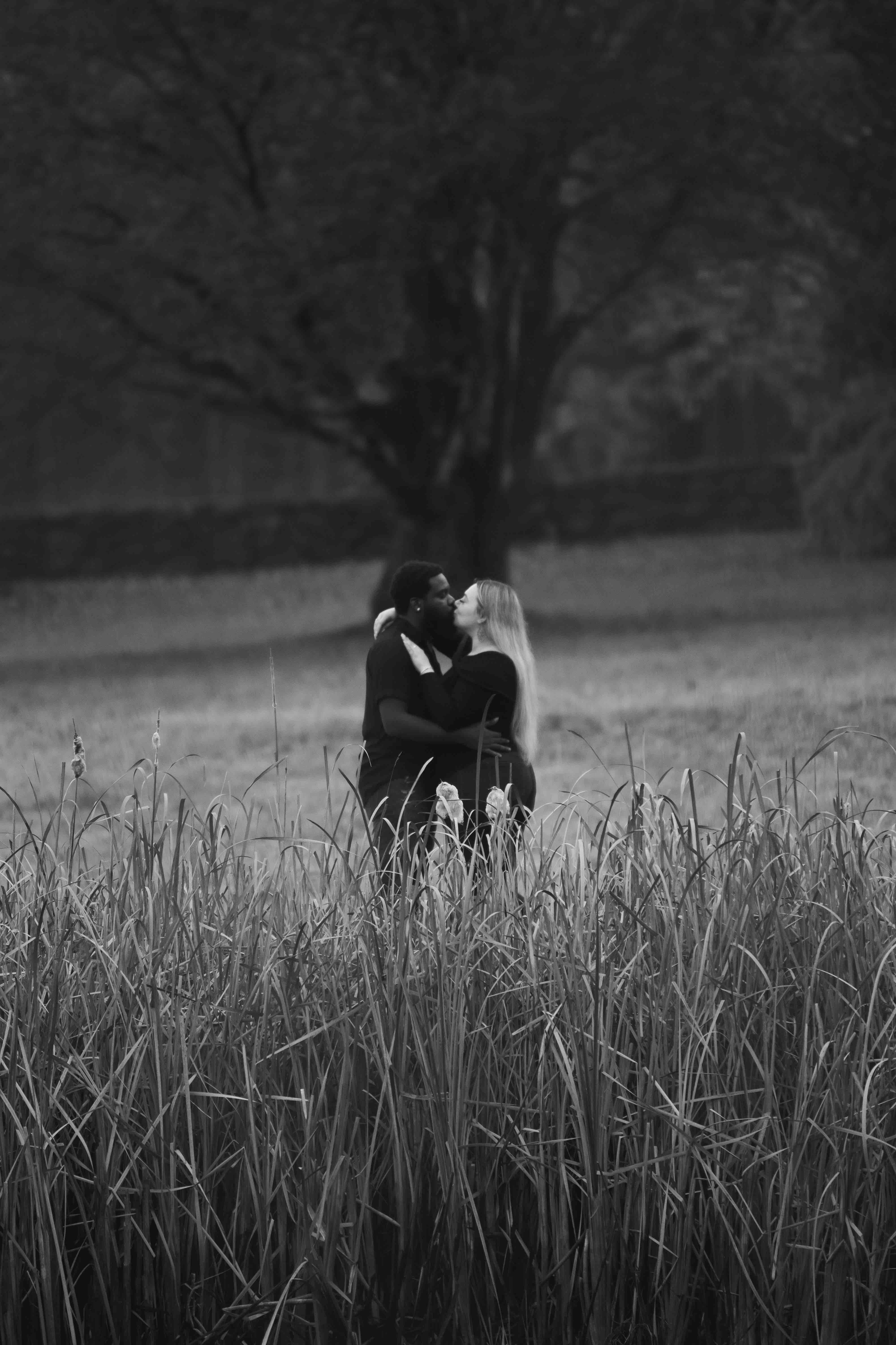 Black and white photo of a couple embracing in a grassy field with trees in the background.