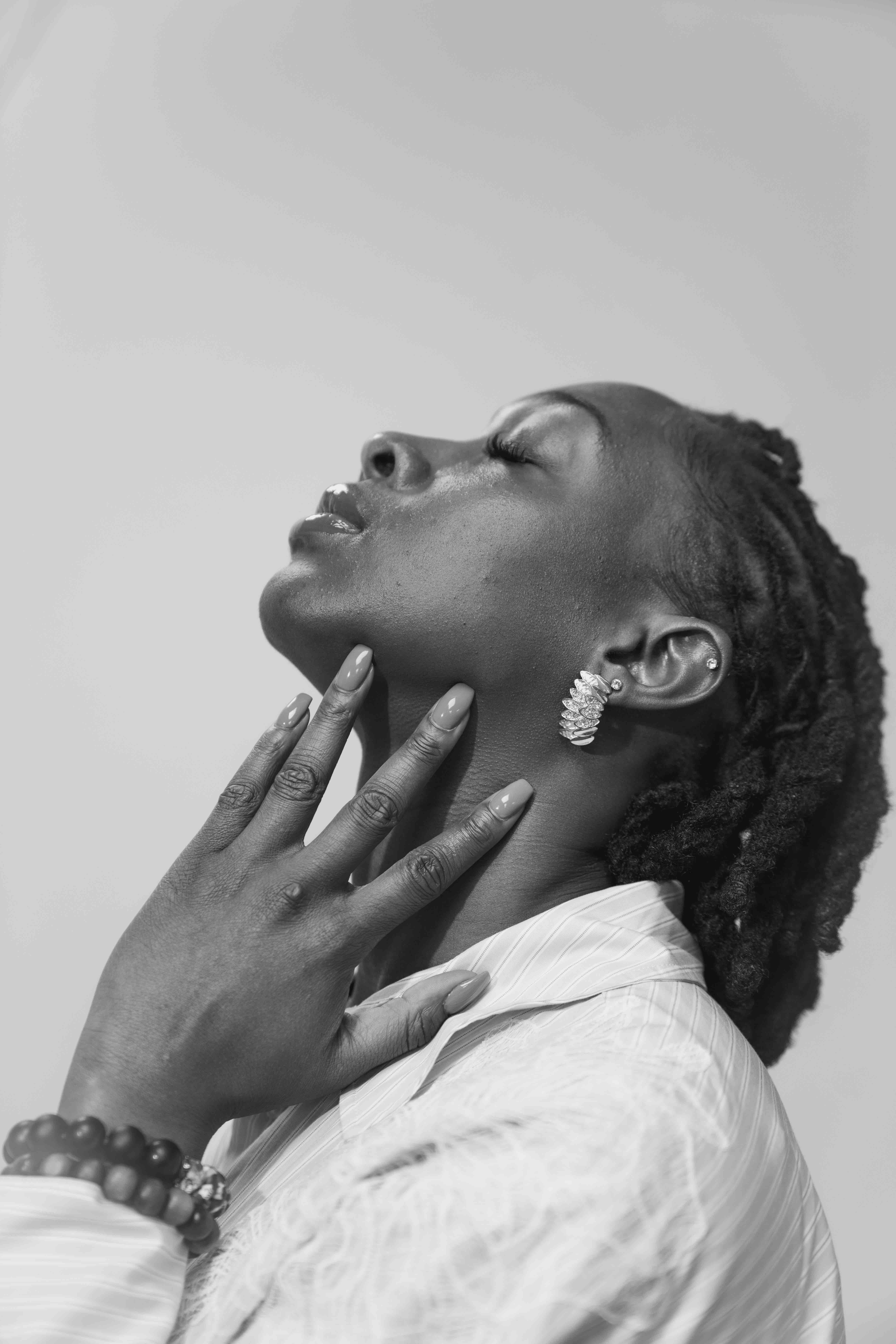 Black woman in profile with eyes closed, hand on neck, wearing earrings, bracelet, and a collared shirt.