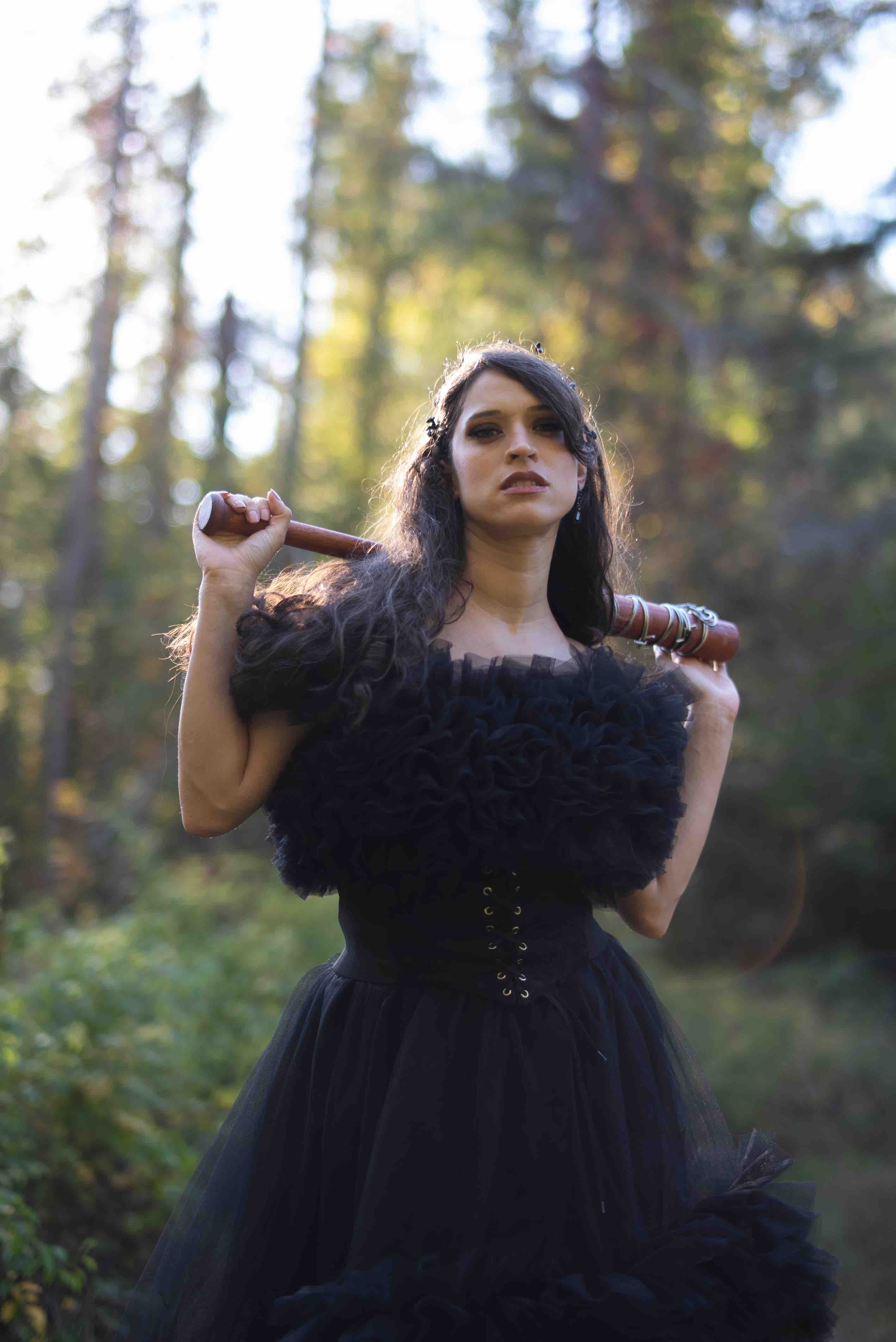 Woman in a black ruffled dress holding a barbed wire wooden bat over her shoulders in a forest.