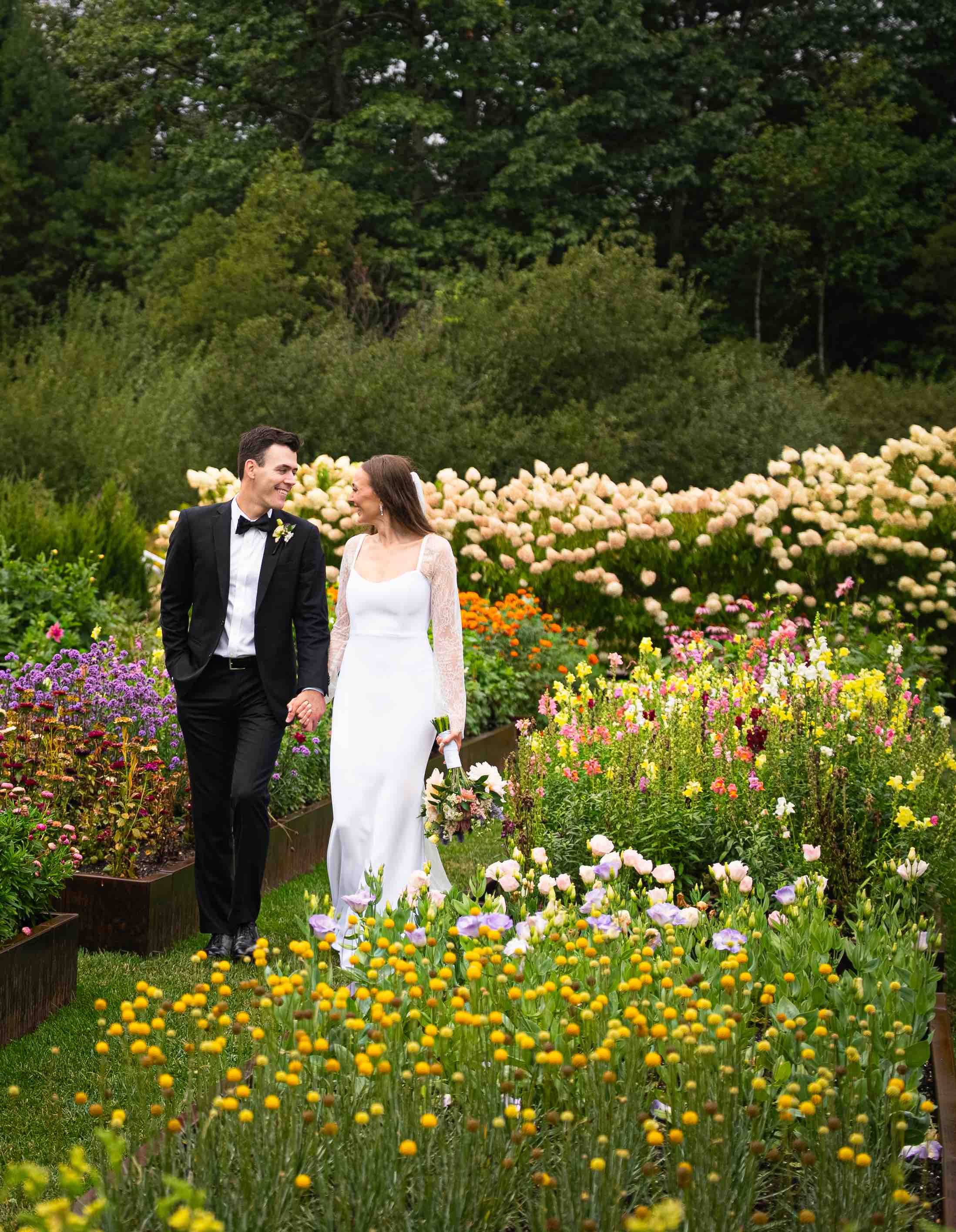 Bride in white gown and groom in black tuxedo holding hands walking through a vibrant garden.