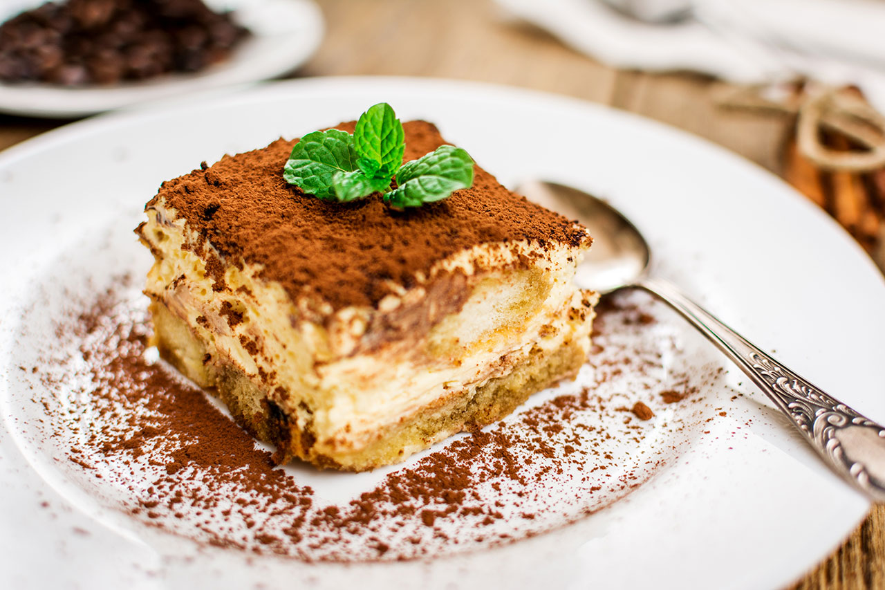 Square slice of tiramisu dusted with cocoa powder and topped with a fresh mint leaf on a white plate with a decorative spoon.