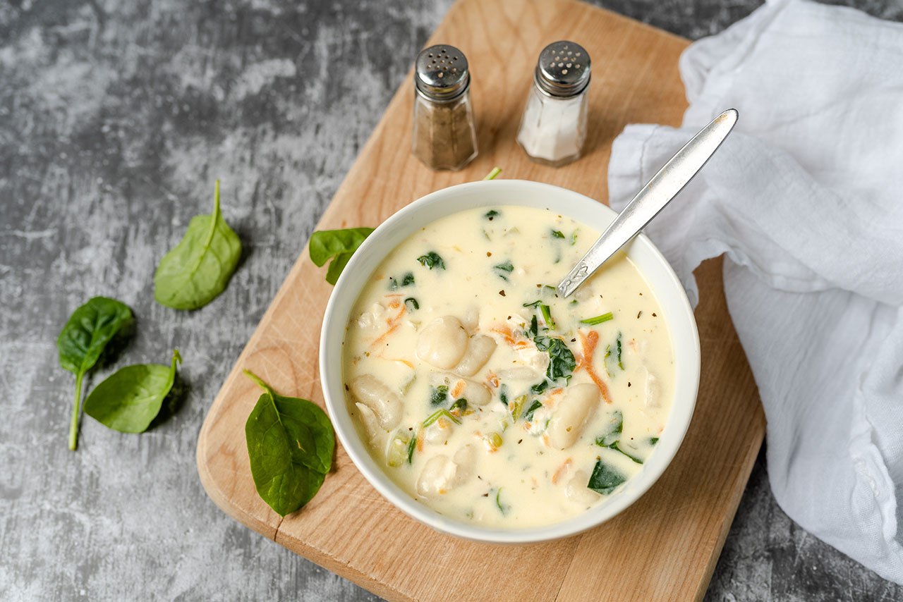 Creamy soup with gnocchi and spinach served in a white bowl on a wooden board with salt and pepper shakers.