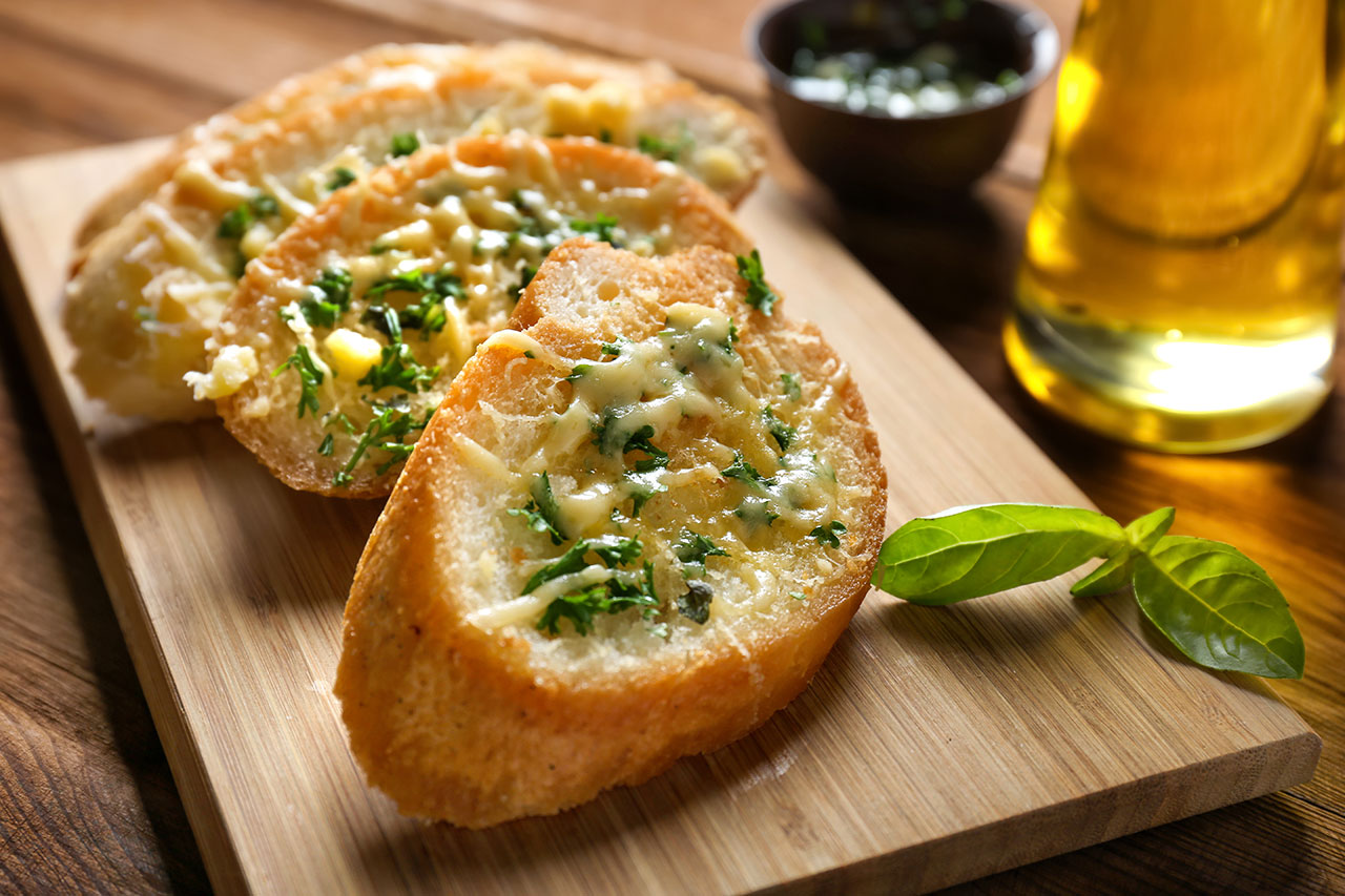 Three slices of garlic bread with melted cheese and chopped parsley on a wooden cutting board, with a basil leaf and a glass of golden liquid in the background.