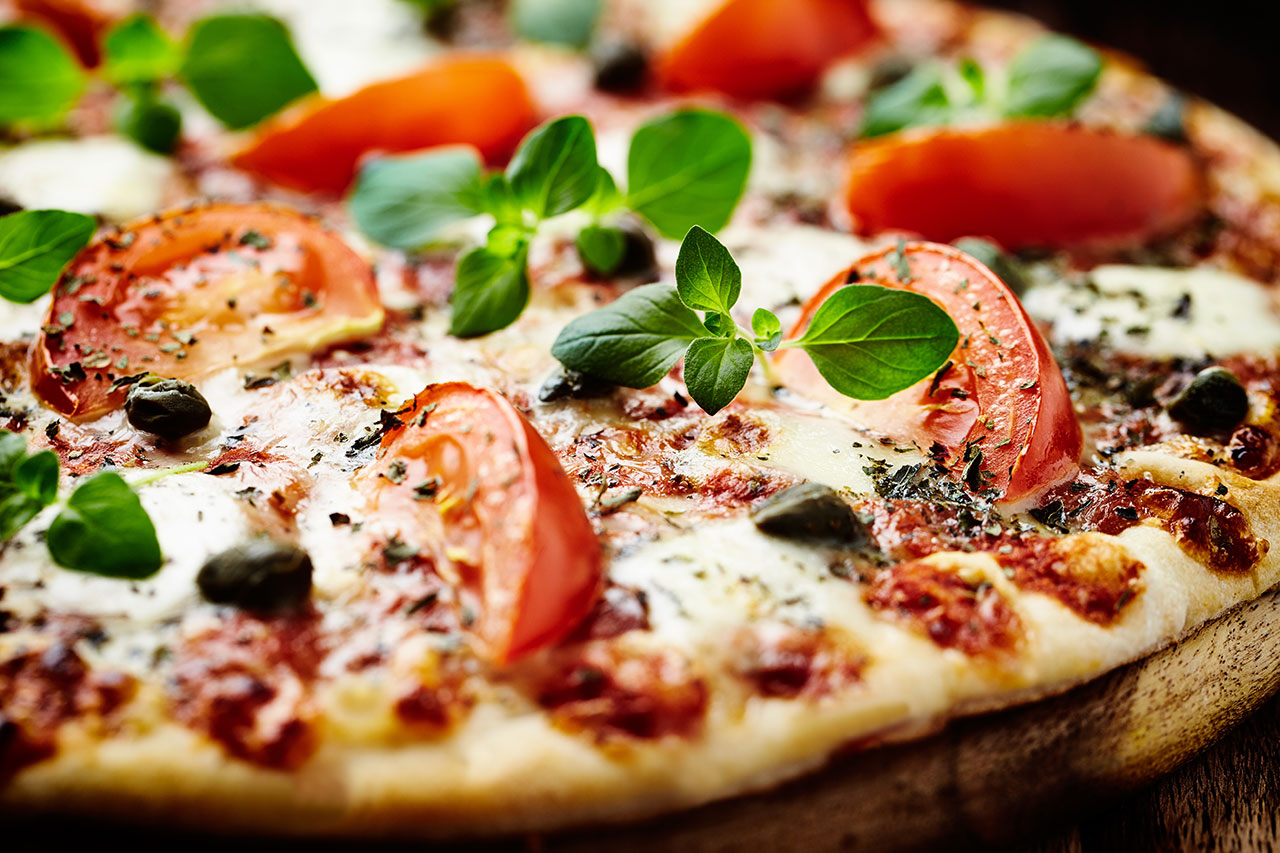 Close-up of Margherita pizza topped with tomato slices, fresh basil leaves, melted mozzarella cheese, and herbs.