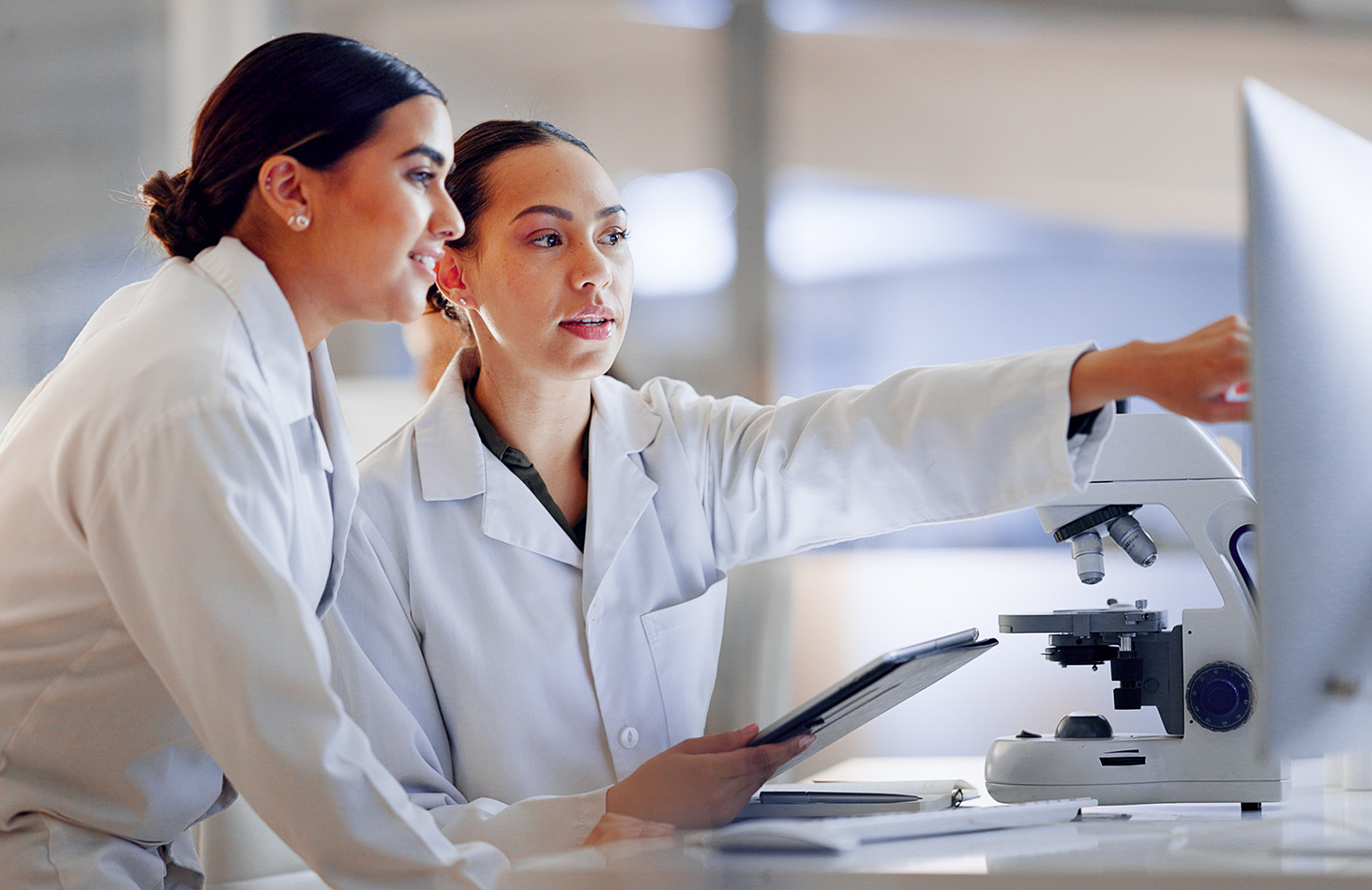 Zwei Wissenschaftlerinnen in weißen Kitteln arbeiten gemeinsam am Mikroskop im Labor.