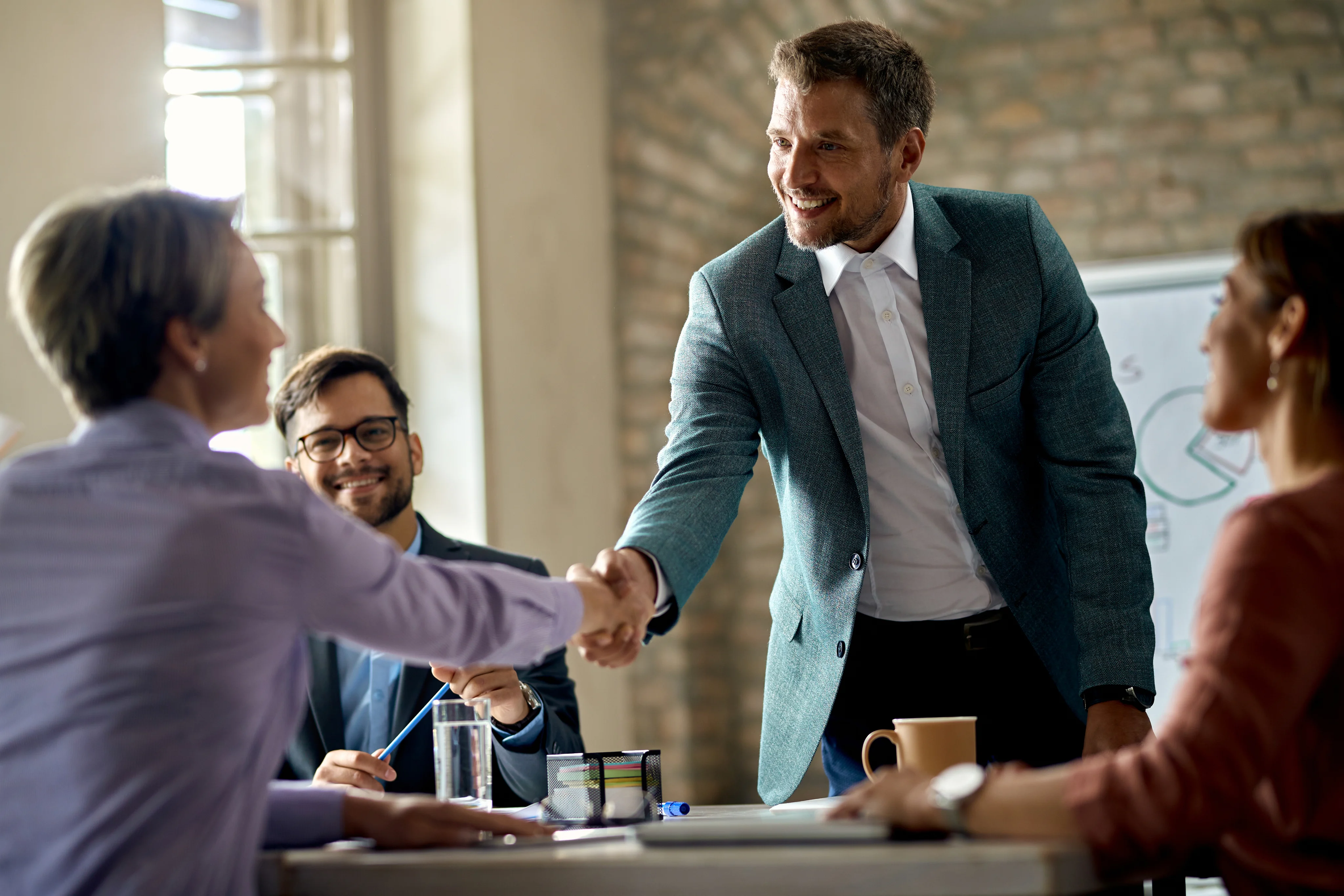 Two businesspeople shaking hands across a table while two colleagues smile and watch during a meeting.